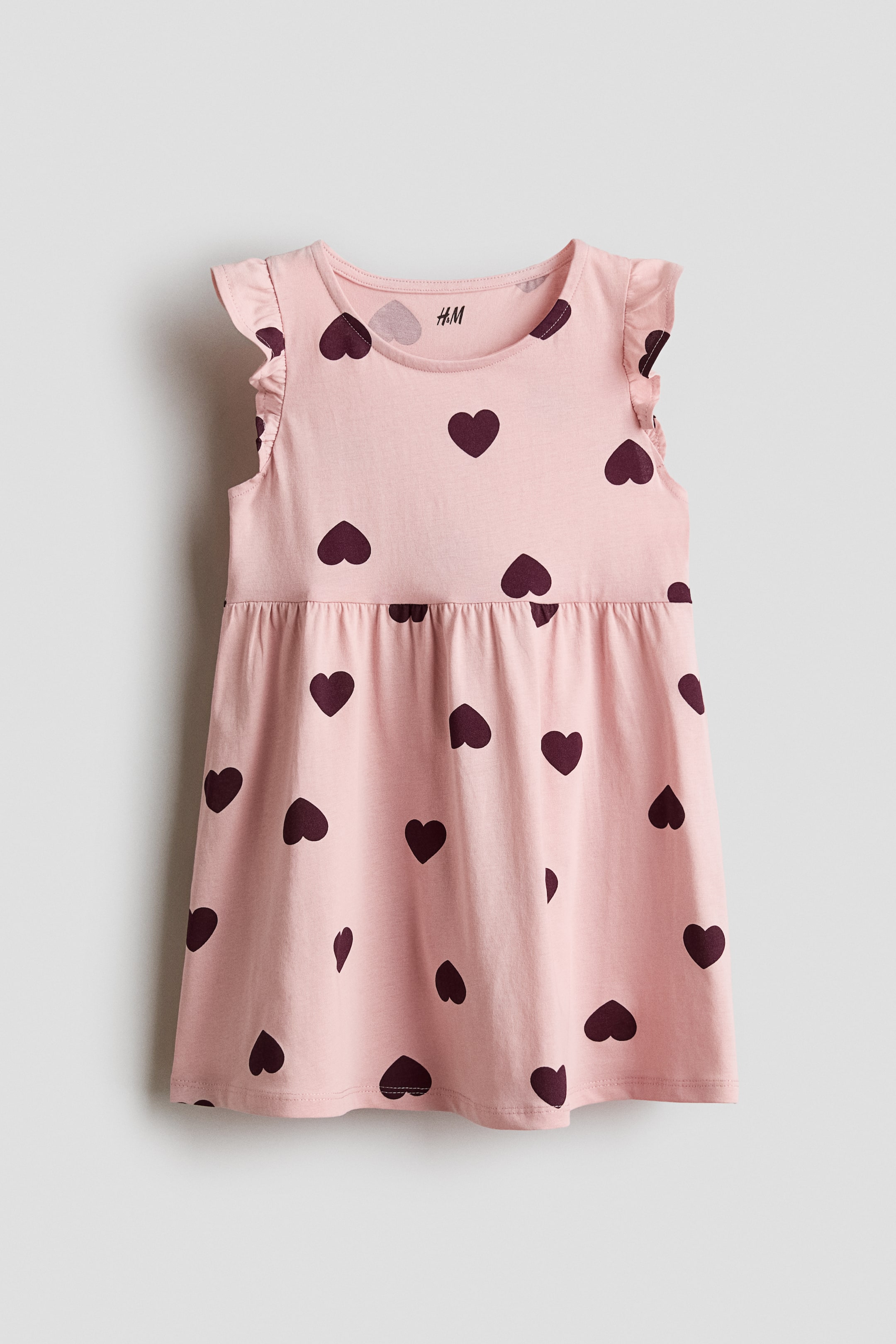 Ampliar la imagen: El frente de un vestido rosa claro muestra un patrón repetido de corazones burdeos oscuros. Está diseñado con un escote redondo, mangas con volantes y una cintura fruncida.