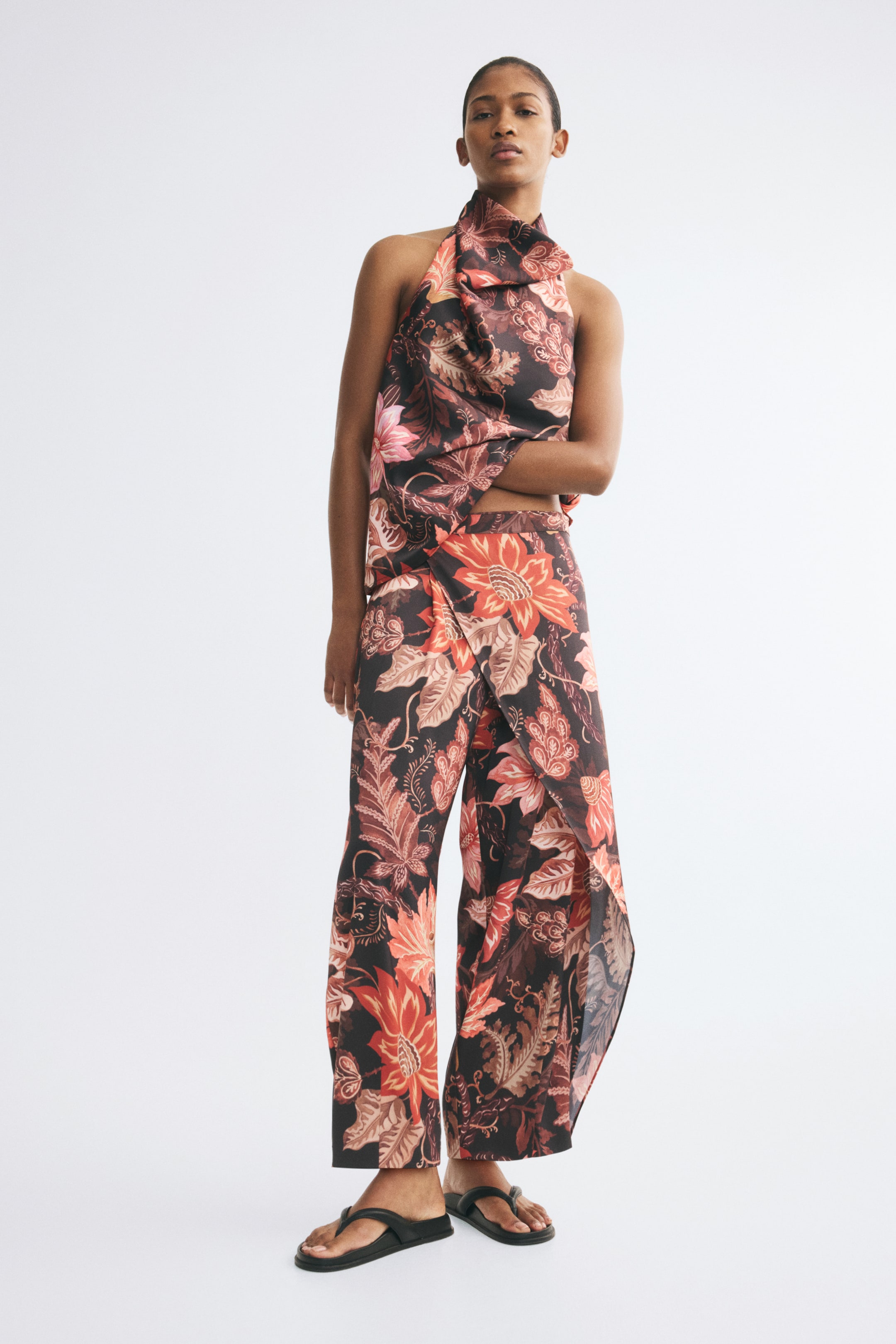 View larger image: Loose-fit satin pants featuring a dark floral print in shades of orange, pink, and brown are being worn by the model, showcasing a draped wrap detail at the front. The wide-leg pants are styled with a matching halter-neck top and black flat sandals.