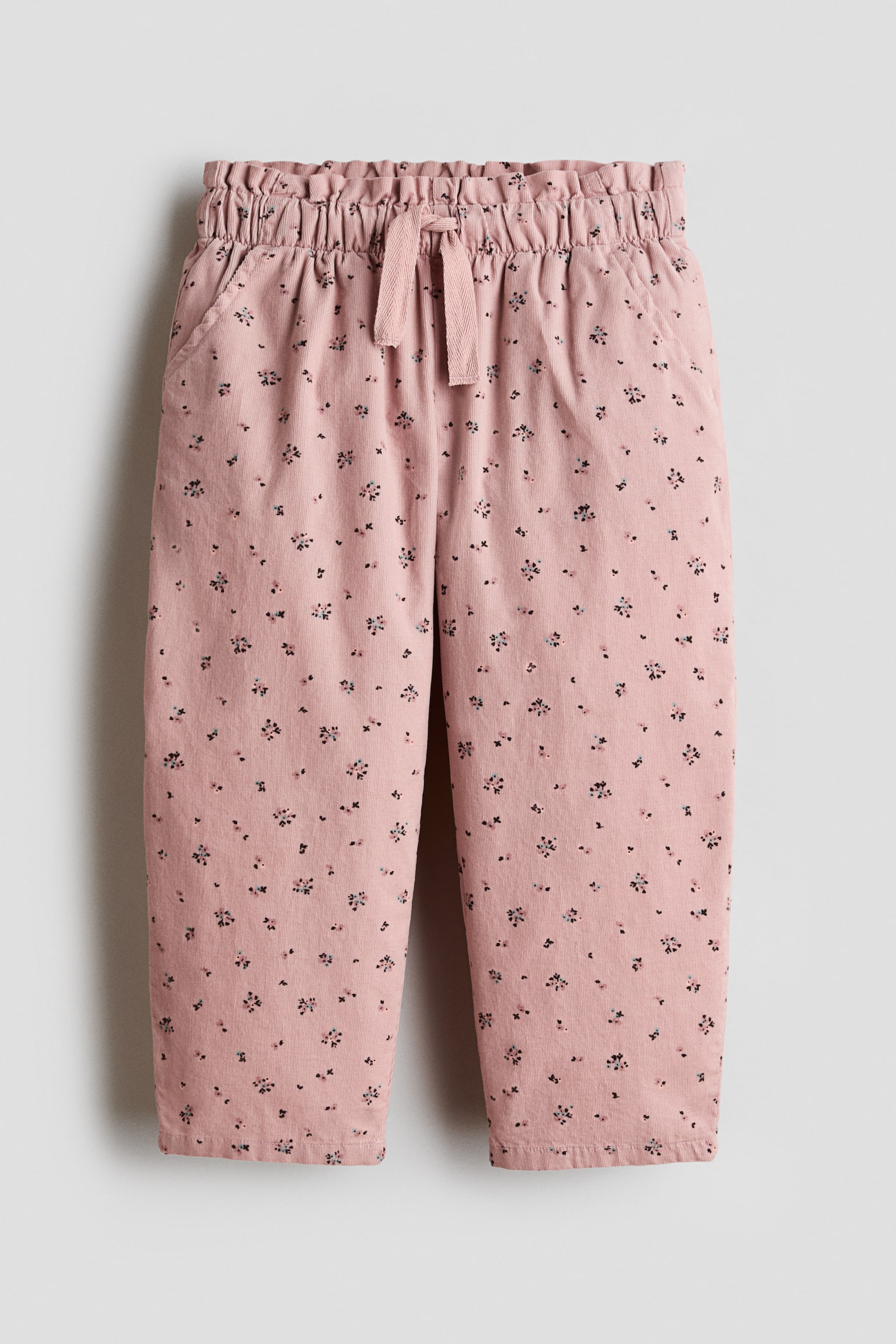 A pair of light pink trousers is shown from the front, showcasing an all-over ditsy floral print and a relaxed straight-leg fit. The trousers feature an elasticated paperbag waistband with a central drawstring tie and side-seam pockets.