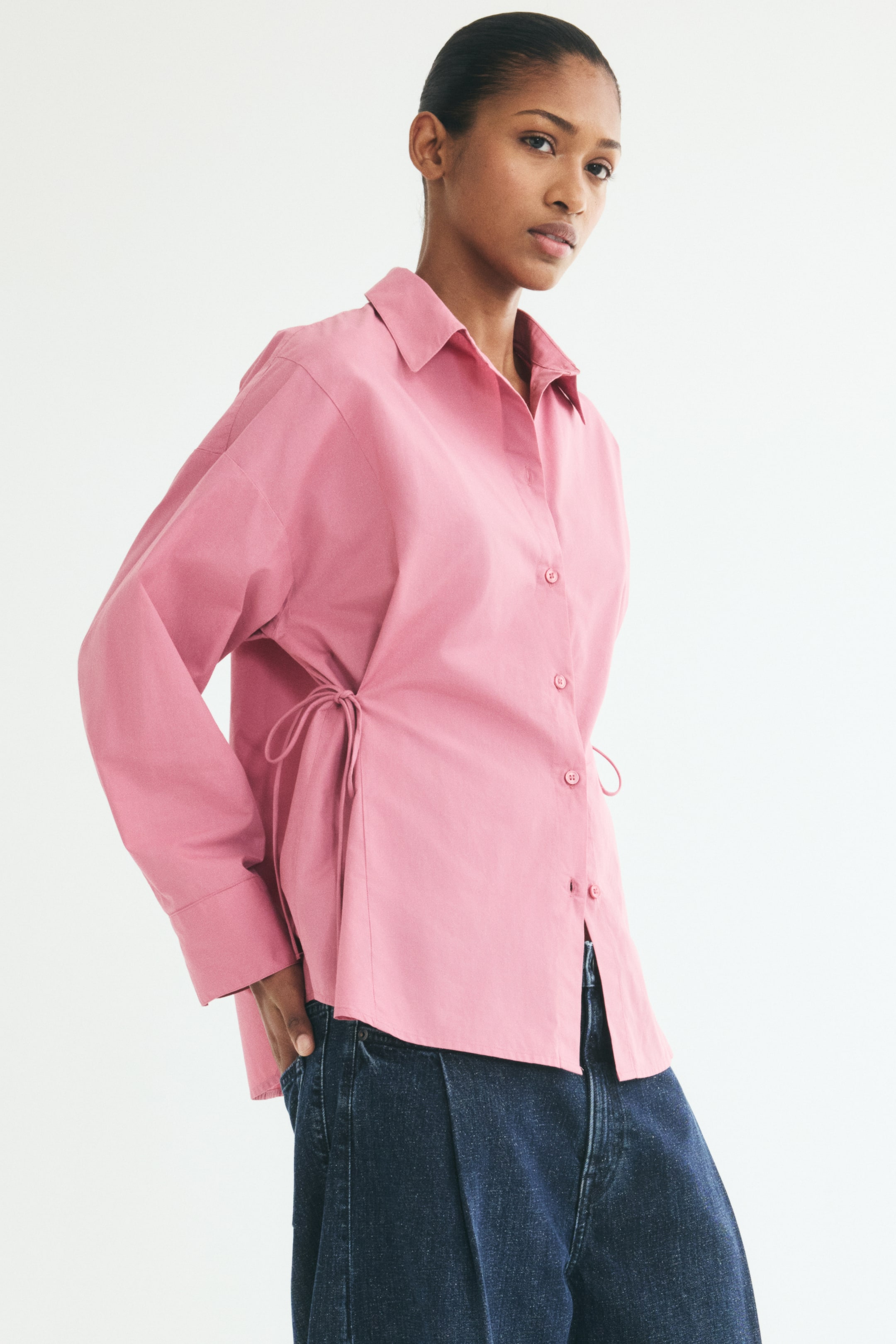 View larger image: A pink woven shirt is shown angled to the right, featuring a collar, button-down front, dropped shoulders, and long sleeves with button cuffs. The shirt is designed with narrow ties on each side of the waist to create a nipped-in effect, with one tie visible on the side.