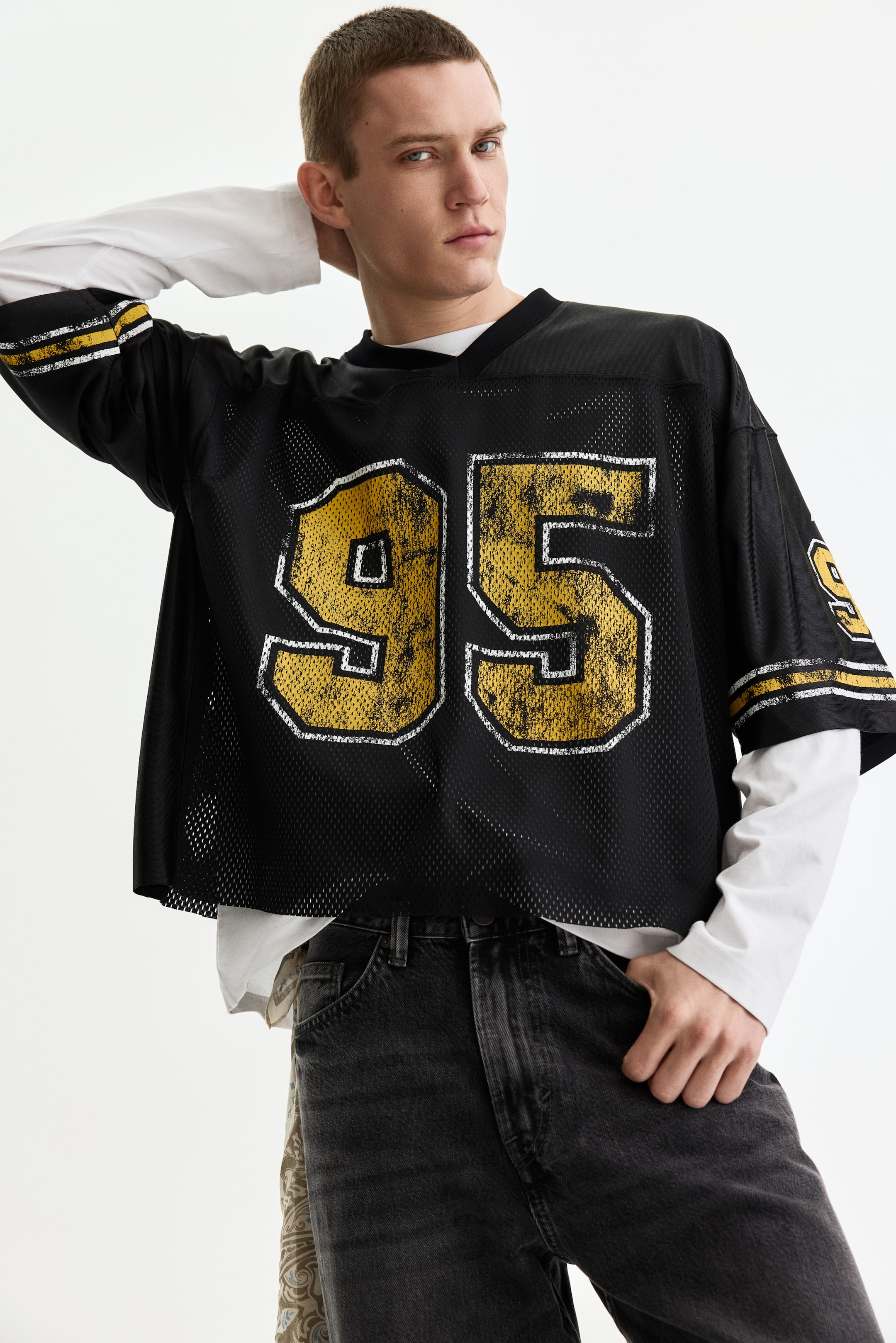 View larger image: A black mesh jersey features a distressed gold-and-white '95' graphic on the front. It has a V-neck, short sleeves with two gold stripes, an oversized cropped fit, and is layered over a white long-sleeve shirt.