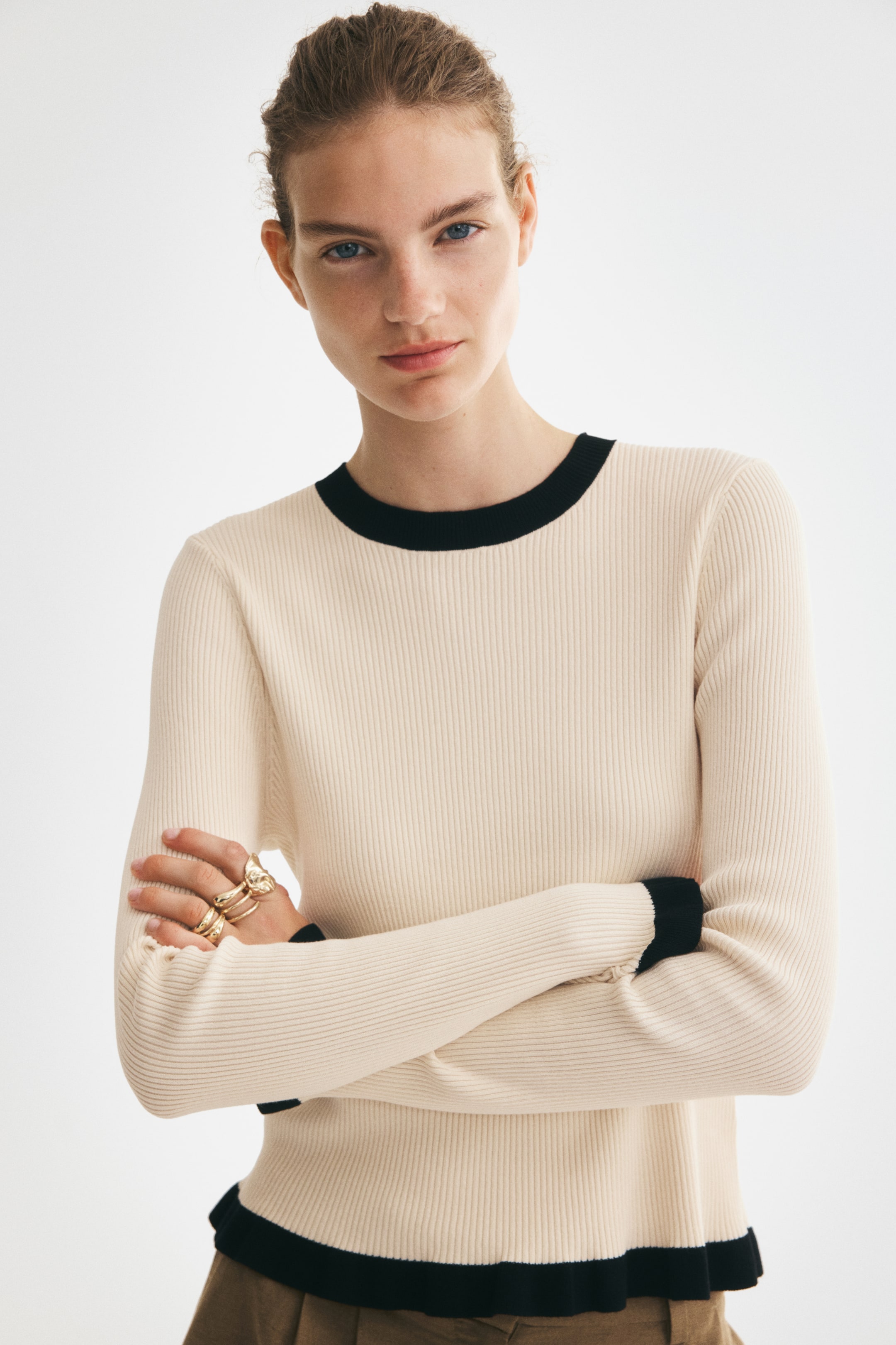 View larger image: A cream-coloured, rib-knit long-sleeve top is being worn by the model, who is standing with arms crossed. The fitted top features a contrasting black round neckline and matching black trims at the cuffs and frilled hem.