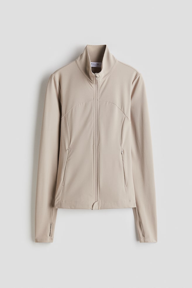A light beige athletic jacket is being presented from the front, featuring a high stand-up collar and a full-length front zipper. It includes contoured seaming on the torso, two angled zippered pockets, and long sleeves with thumbholes at the cuffs. - 1