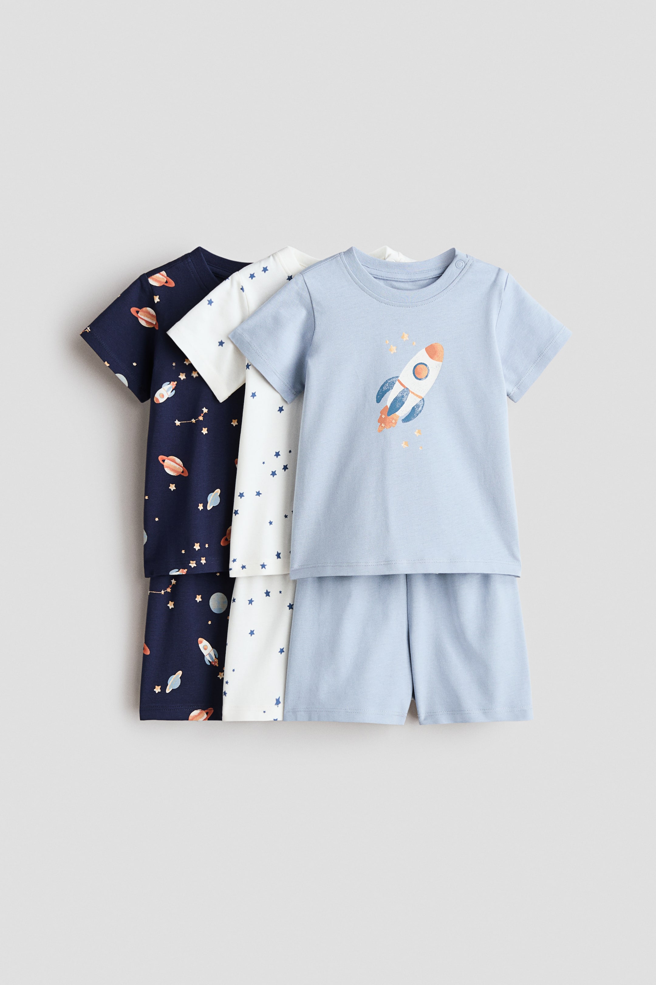 이미지 확대 보기: 세트로 구성된 세 가지 짧은 면 파자마가 앞면을 보여주며 약간 겹쳐 쌓여 있습니다. 가장 앞에 있는 세트는 라이트 블루 색상으로, 로켓 그래픽이 있는 반팔 상의와 매칭되는 반바지로 구성되어 있습니다. 중간 세트는 흰색이며, 반팔 상의와 반바지에 전체적으로 파란 별 패턴이 있습니다. 가장 뒤에 있는 세트는 네이비 블루 색상으로, 반팔 상의와 반바지에 전체적으로 우주 테마의 패턴이 있습니다.