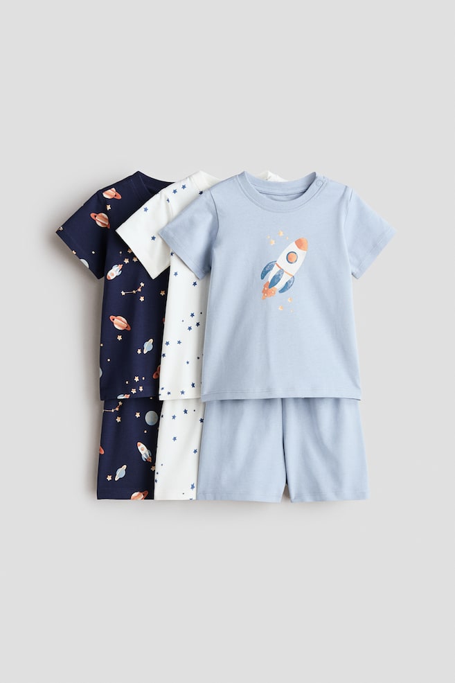 Trois ensembles de pyjamas courts en coton sont présentés de face, empilés légèrement décalés. L'ensemble le plus à l'avant est bleu clair avec un haut à manches courtes arborant un graphique de fusée et un short assorti ; l'ensemble du milieu est blanc avec un motif intégral d'étoiles bleues sur le haut à manches courtes et le short ; et l'ensemble le plus à l'arrière est bleu marine avec un motif intégral sur le thème de l'espace sur le haut à manches courtes et le short. - 1