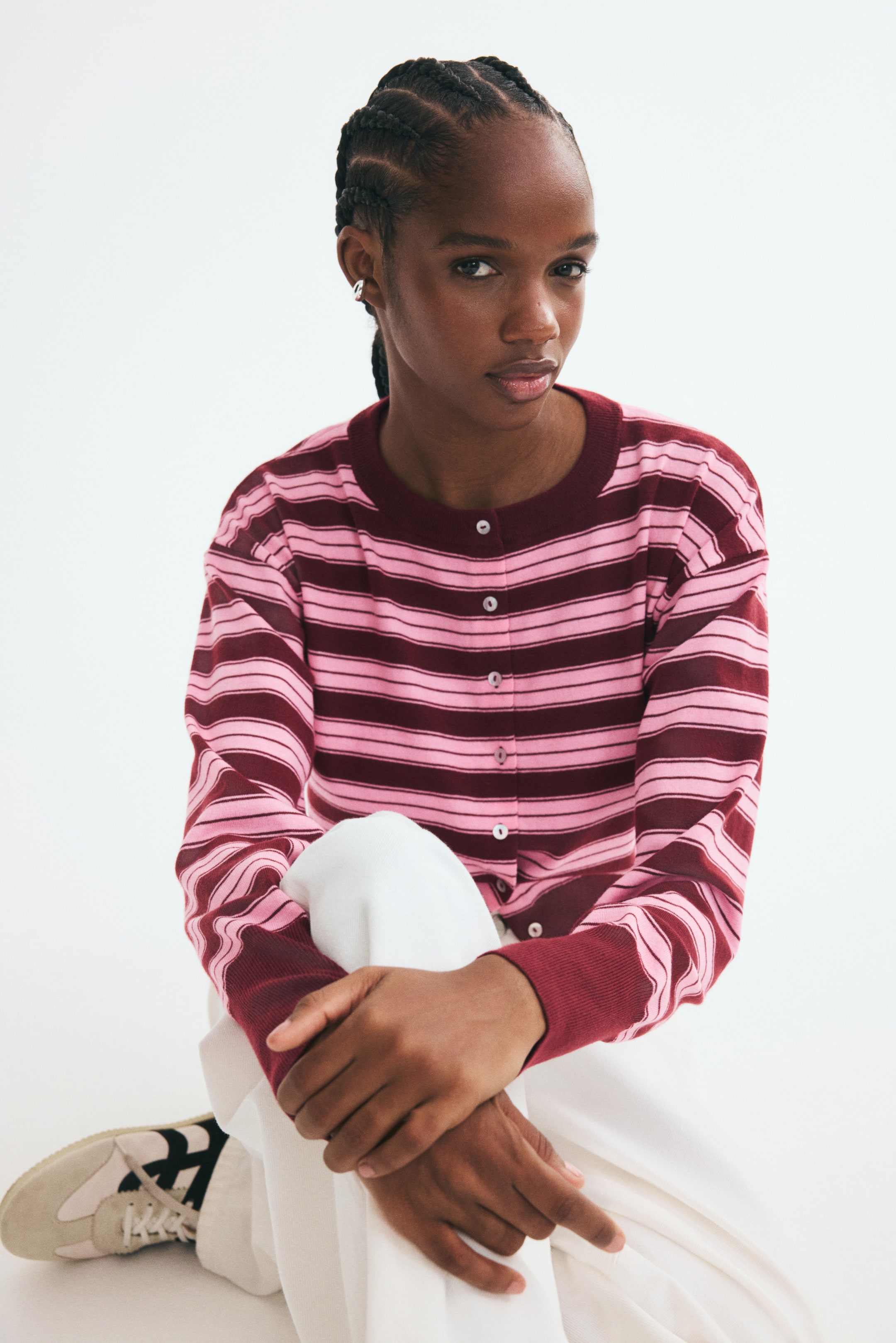 View larger image: A short, fine-knit cardigan with pink and burgundy horizontal stripes is worn by the model, featuring a ribbed round neckline, button-front closure, dropped shoulders, and wide burgundy cuffs. The cardigan is styled with white trousers as the model sits with hands clasped over one knee.