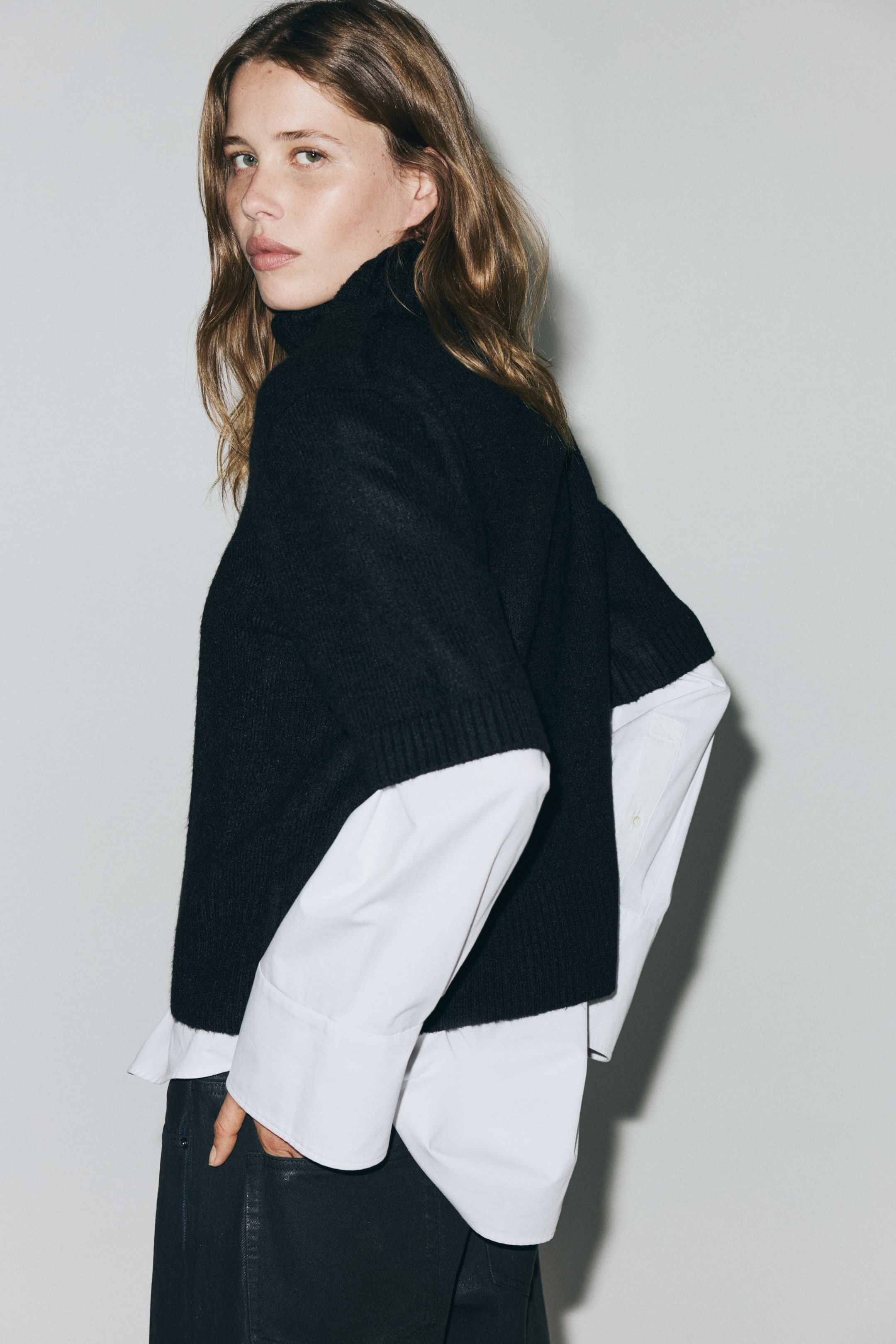 View larger image: A black short-sleeved turtleneck sweater is being presented from a back-side angle, layered over a white long-sleeved shirt. The sweater is featuring a ribbed turtleneck and ribbed short sleeves, and the white shirt's long cuffs are extending past its hem.