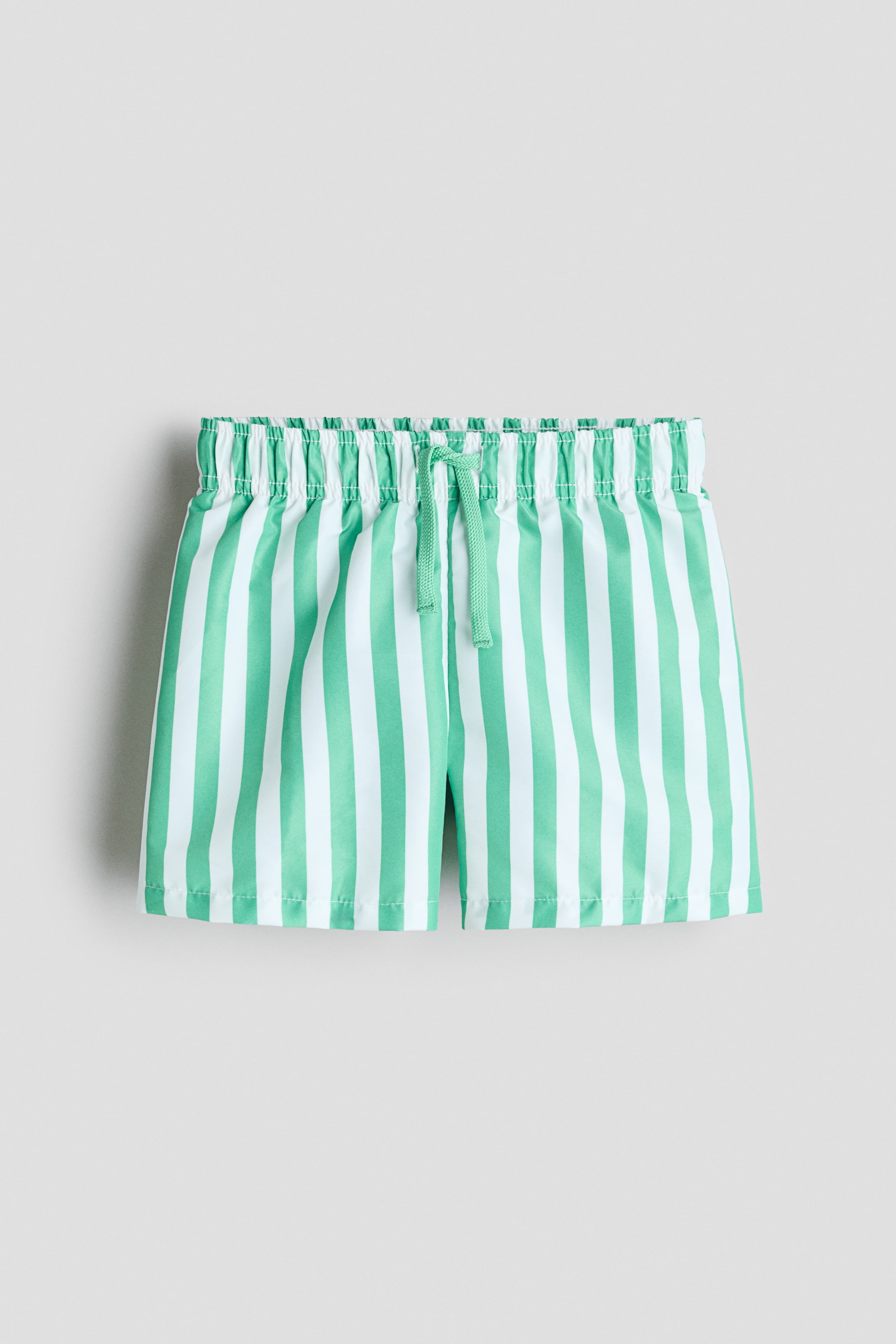 View larger image: These printed swim shorts showcase green and white vertical stripes. They feature an elastic waist with a green drawstring tied at the front.