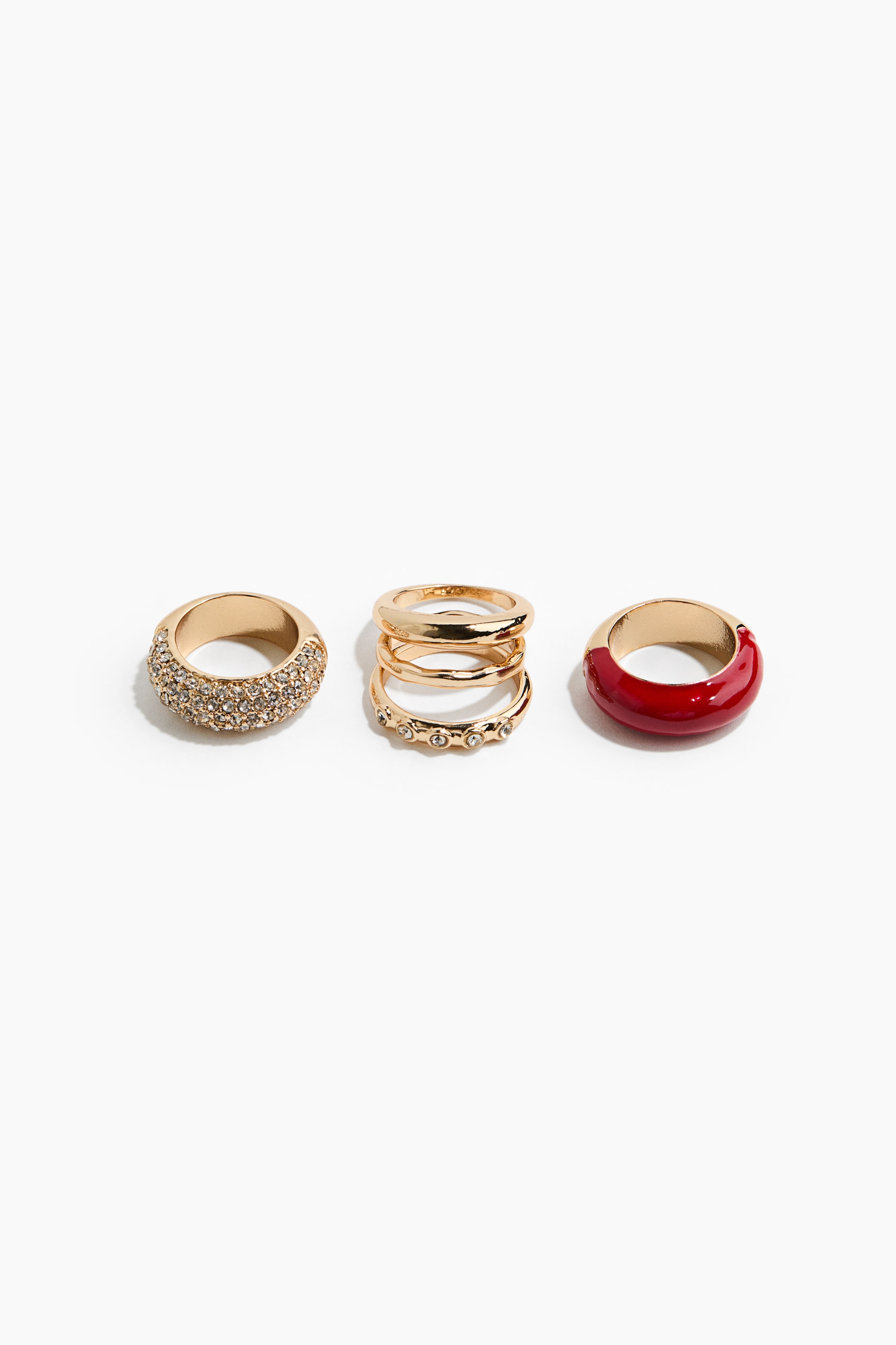 View larger image: A set of gold-toned rings presents three distinct styles arranged side by side. The set includes a wide band fully embellished with clear rhinestones, a stack of three thin gold-toned bands with the bottom one featuring small inset crystals, and a wide band with a glossy red enamel coating.