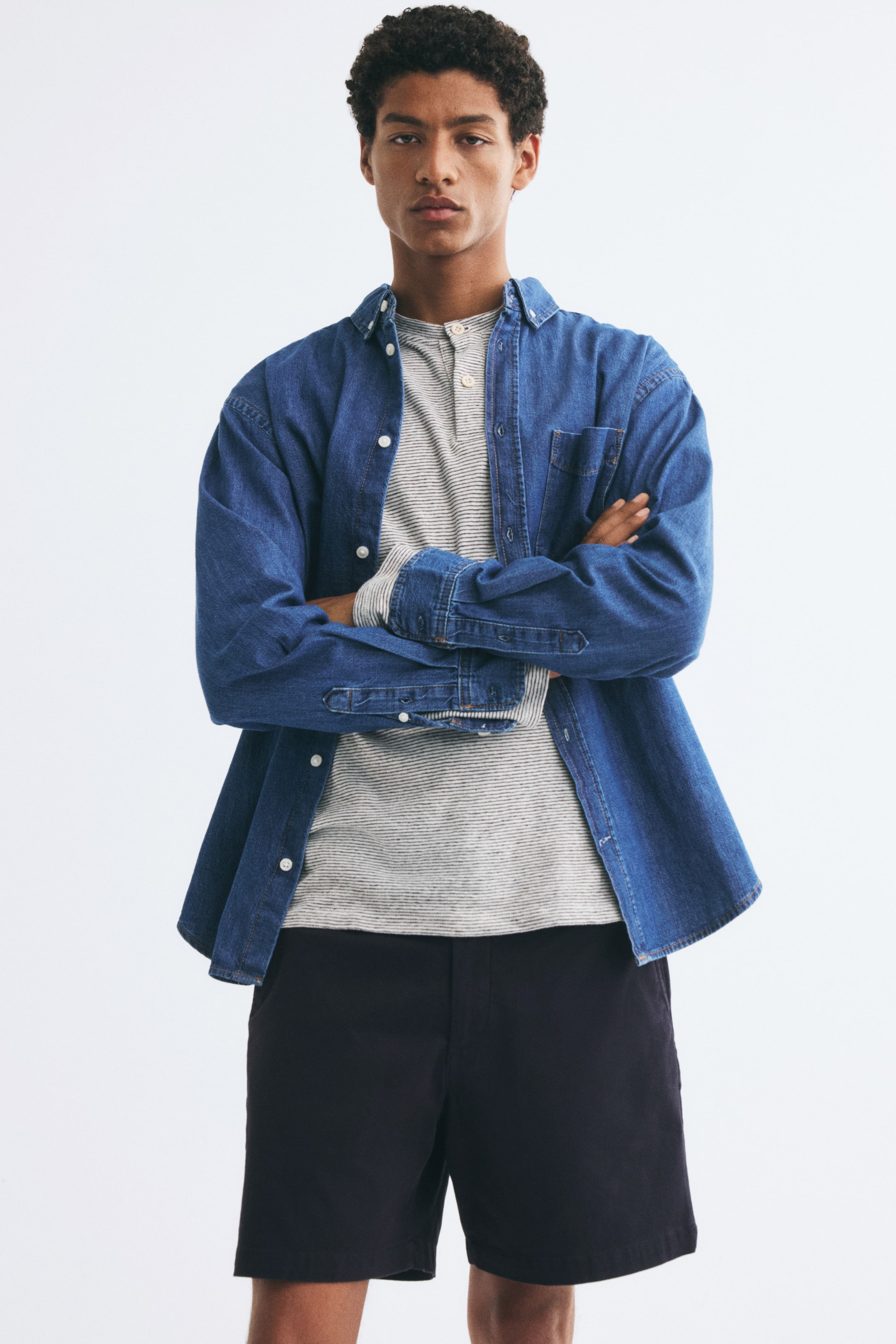 View larger image: The model is wearing dark chino shorts in a regular fit, styled with a striped henley shirt and an open denim shirt. The shorts feature a classic silhouette and fall just above the knee.