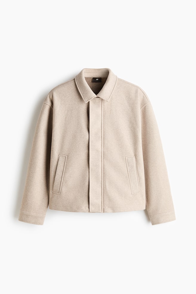 Ein hellbeiges Overshirt aus weichem, filzartigem Stoff hÃ¤ngt frontseitig und zeigt einen klassischen Kragen sowie eine verdeckte FrontverschlieÃung. Die Jacke verfÃ¼gt Ã¼ber Ã¼berschnittene Schultern, lange Ãrmel, seitliche Leistentaschen und einen geradlinigen Saum. - 1