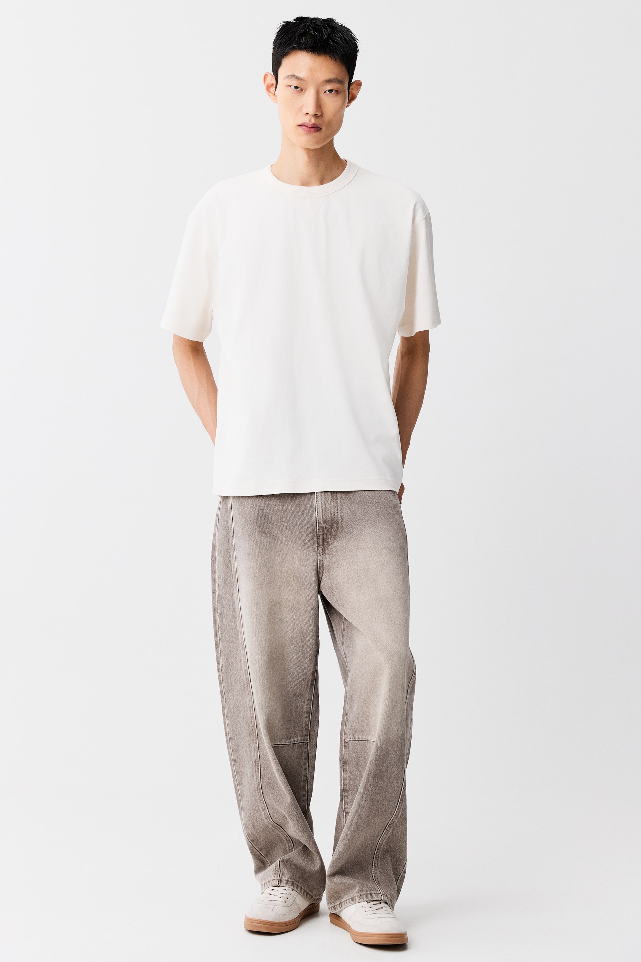 拡大画像を表示: モデルは、リラックスフィットのオフホワイトのクルーネックTシャツを着用し、目立つシームディテールが施されたワイドレッグのウォッシュドブラウンジーンズ、ガムソールのライトカラーのローカットスニーカーでスタイリングされています。