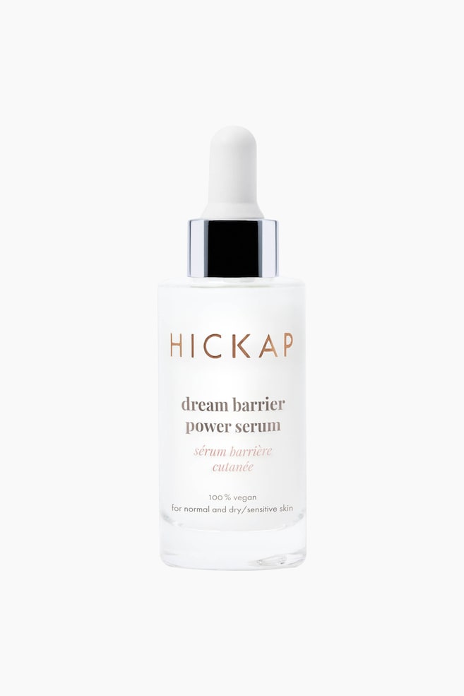 En rektangulär, frostad klar glasflaska som innehåller ett hudvårdsserum står upprätt och vänd framåt. Den har en vit pipettapplikator med en metallisk silverkrage, och dess framsida visar text inklusive 'HICKAP' och 'dream barrier power serum' i en roséguldfärg. - 1