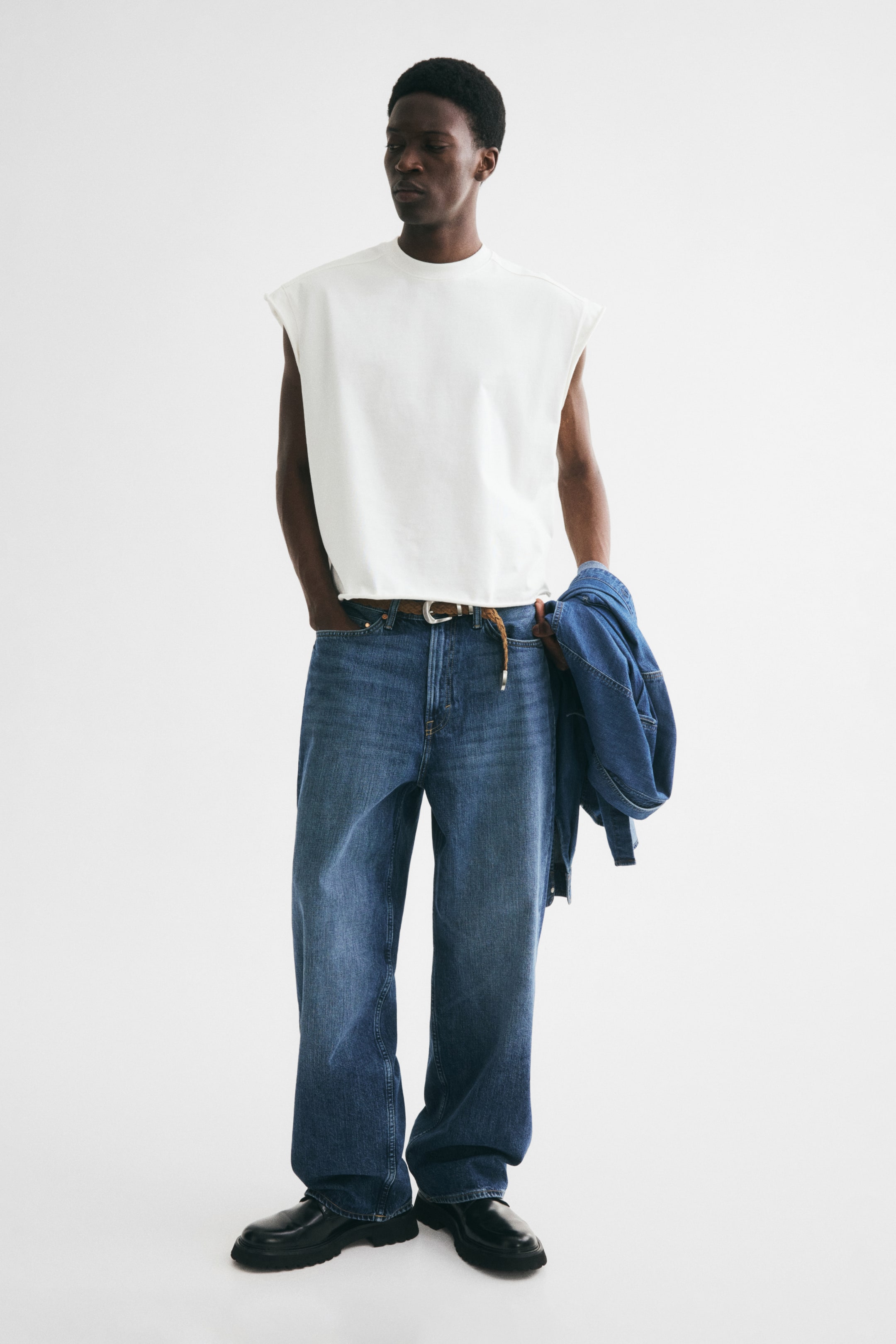 View larger image: Ang modelo ay nakasuot ng asul na denim na 5-pocket jeans na gawa sa matibay na koton. Ang mga ito ay may regular na baywang na may kasamang tinirintas na sinturon, maluwag na fit na may bilugan na binti at bumabagsak na pundya, at nakasalansan sa bukong-bukong sa itaas ng itim na sapatos. Ang kanang kamay ng modelo ay nakapatong sa isang bulsa sa harap, at isang denim na jacket ang hawak sa kaliwang kamay laban sa pantalon.