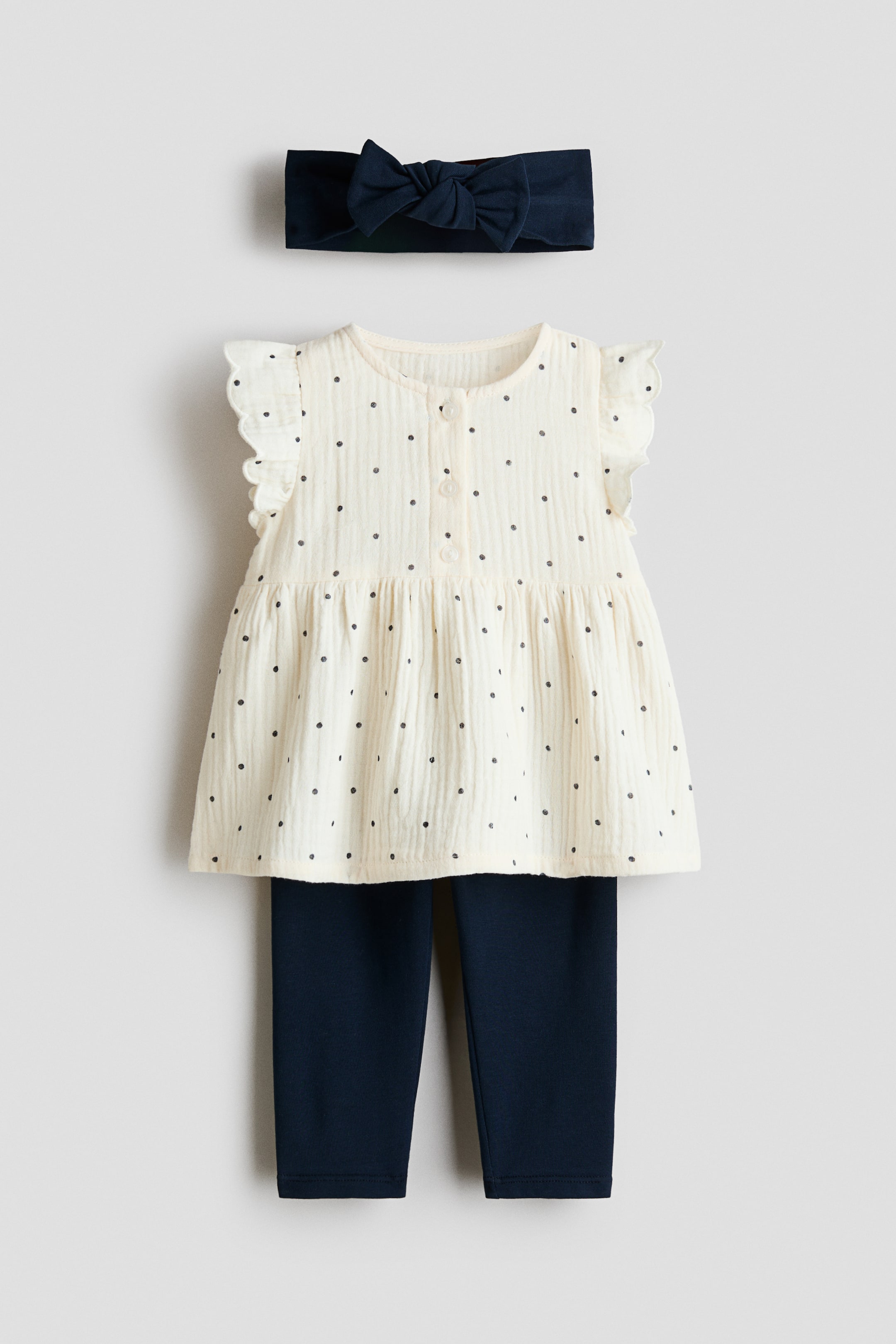 View larger image: A three-piece baby outfit is showcased front-facing, featuring a navy blue bow headband, a cream textured top, and navy blue leggings. The top is styled with small dark polka dots, ruffled cap sleeves, a button placket, and a gathered waist that creates a flared hem.