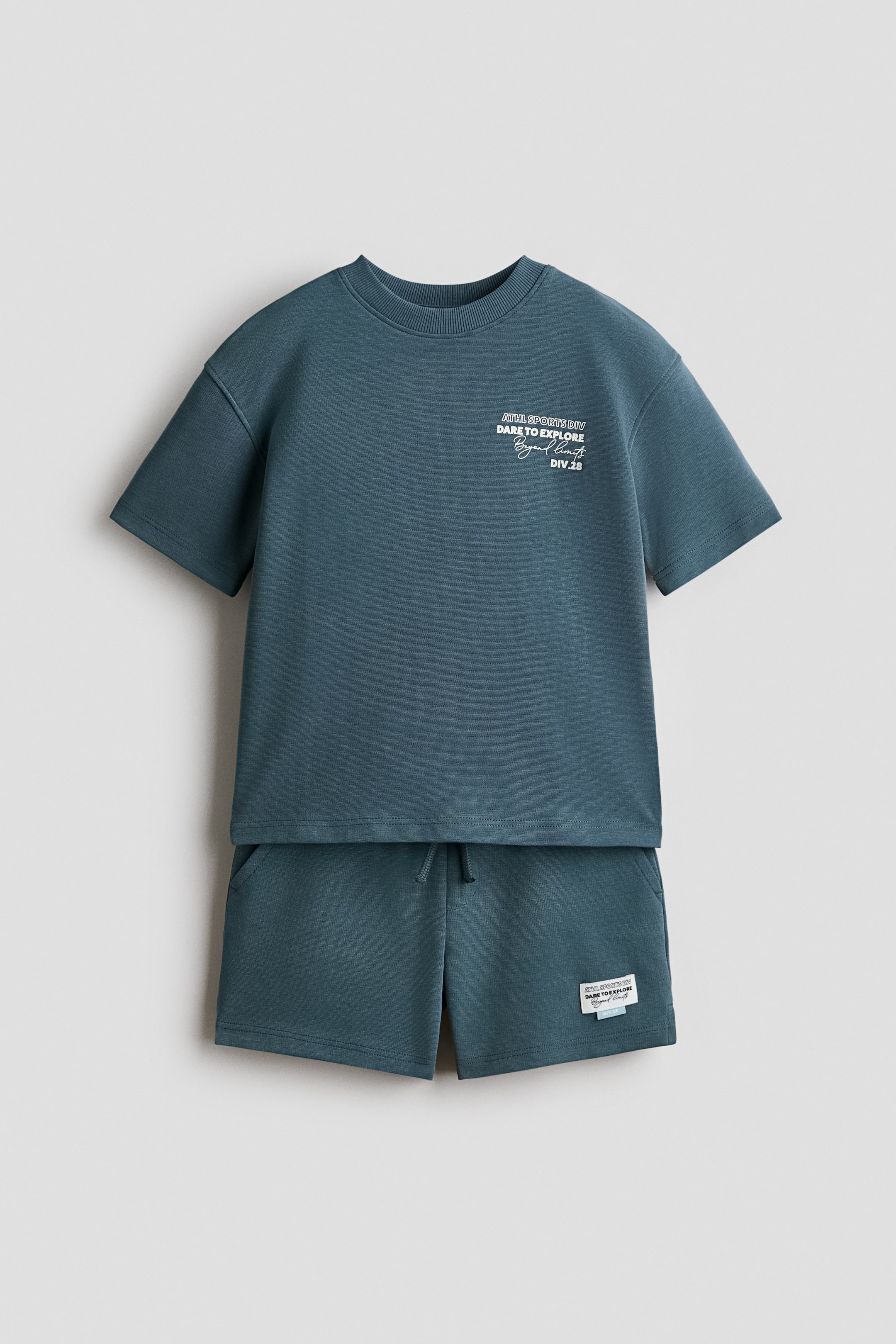 View larger image: A front-facing dark teal set features a T-shirt with a ribbed neckline, gently dropped shoulders, and a white chest graphic, paired with matching shorts. The shorts display an elasticated drawstring waist, a faux fly, diagonal side pockets, and a small white patch on the right thigh.