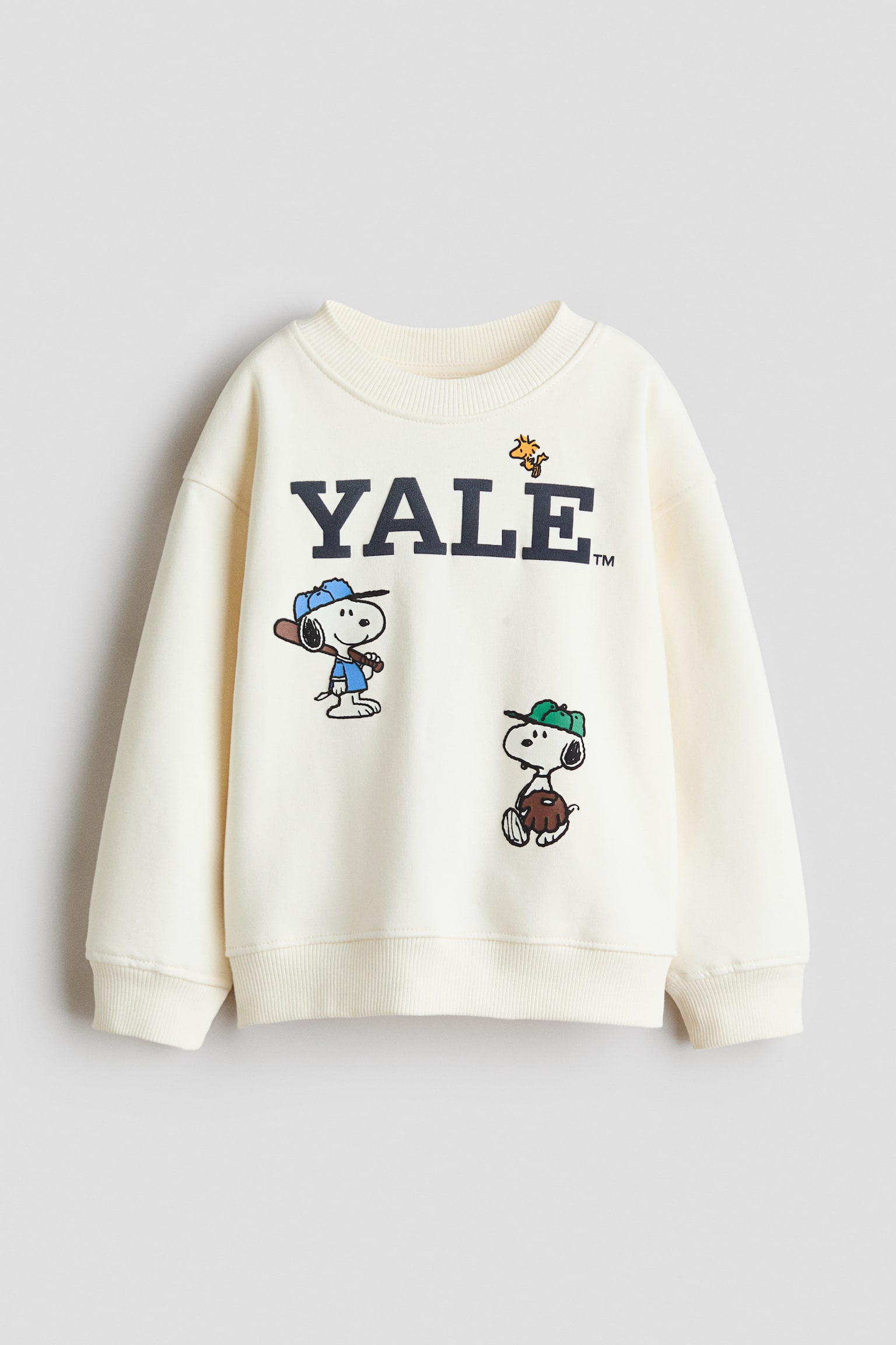 A front view of an off-white, loose-fit sweater featuring a round, rib-trimmed neckline, dropped shoulders, and ribbing at the cuffs and hem. The front showcases a printed design with the word "YALE" in navy block letters, accompanied by graphics of Snoopy and Woodstock dressed in baseball gear.