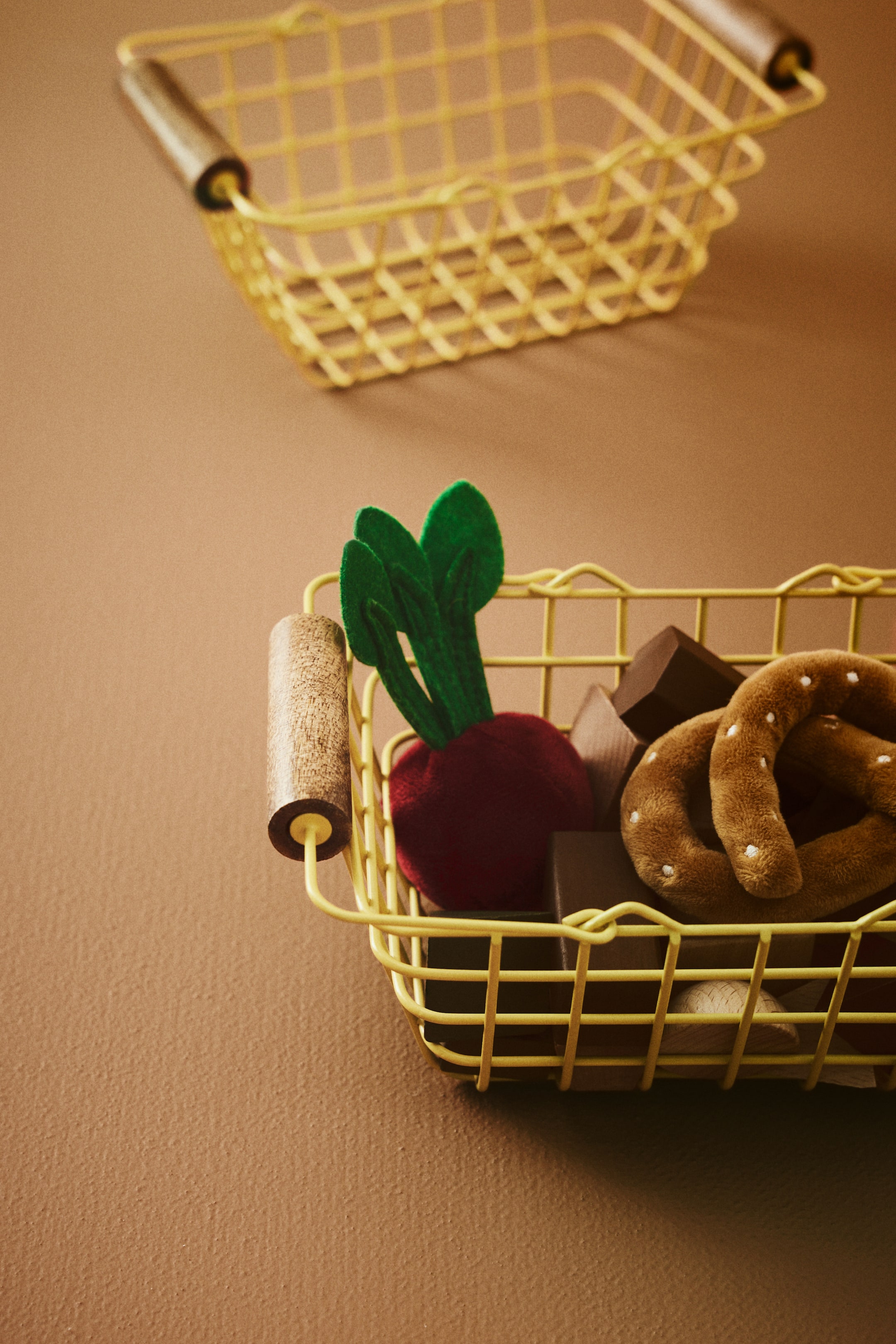 Nagyobb kép megtekintése: Egy téglalap alakú sárga drót kosár tart egy piros plüss céklát zöld filc levelekkel, egy barna plüss perecet fehér pöttyökkel, és sötétbarna fa kockákat. A kosár világosbarna hengeres fogantyúkkal rendelkezik, amelyek mindkét oldalon sötét végekkel vannak ellátva, és kissé megemelt szögből van fényképezve.