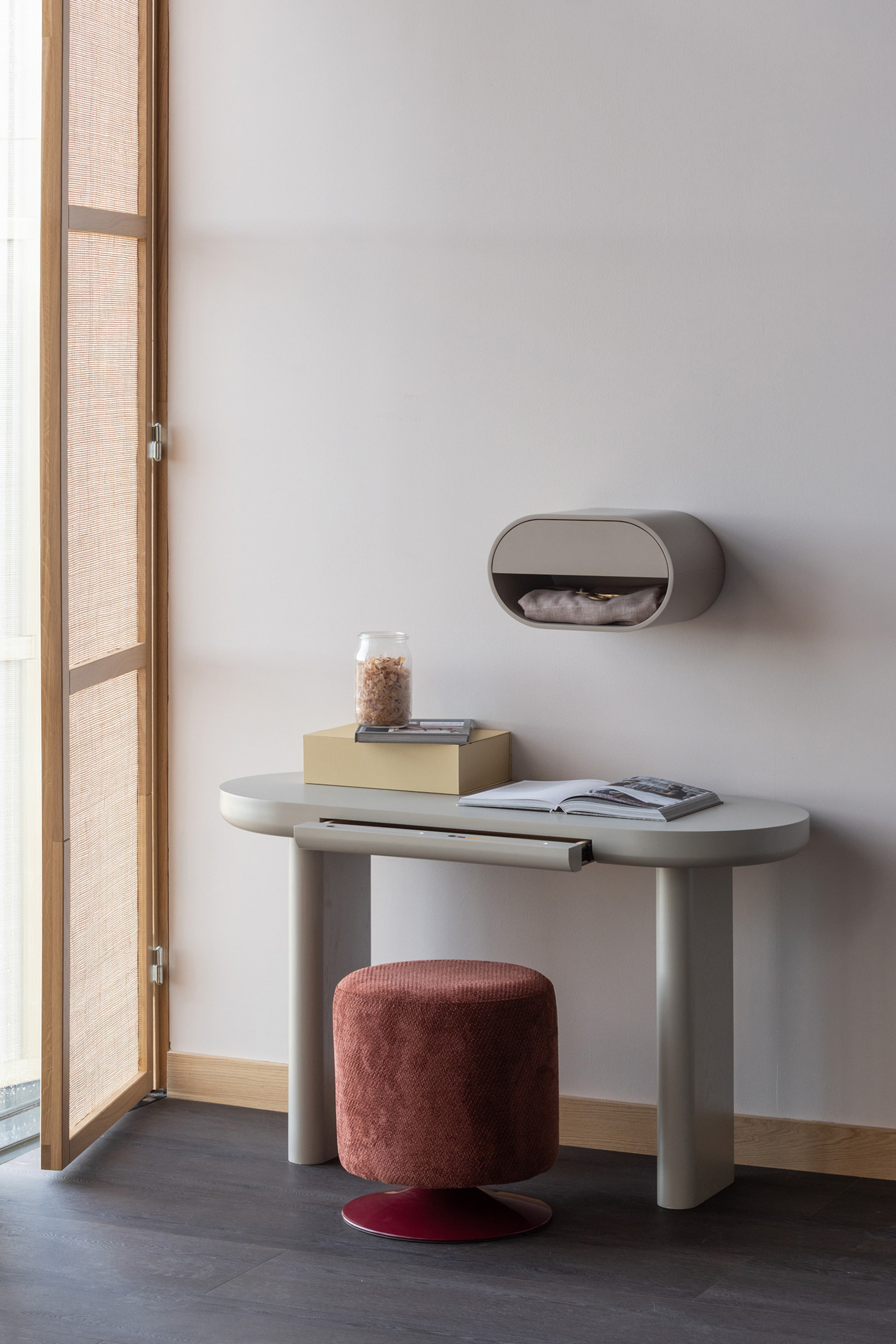 Grotere afbeelding bekijken: Een lichtgrijze console tafel met een ovaal blad en twee dikke, gebogen poten toont zijn voorste rechterzijde, met een ondiepe uitschuiflade die onder het oppervlak vandaan komt. Een bijpassende ovale wandplank is erboven gepositioneerd, met een open compartiment met een schuifpaneel en bevat een opgevouwen item.