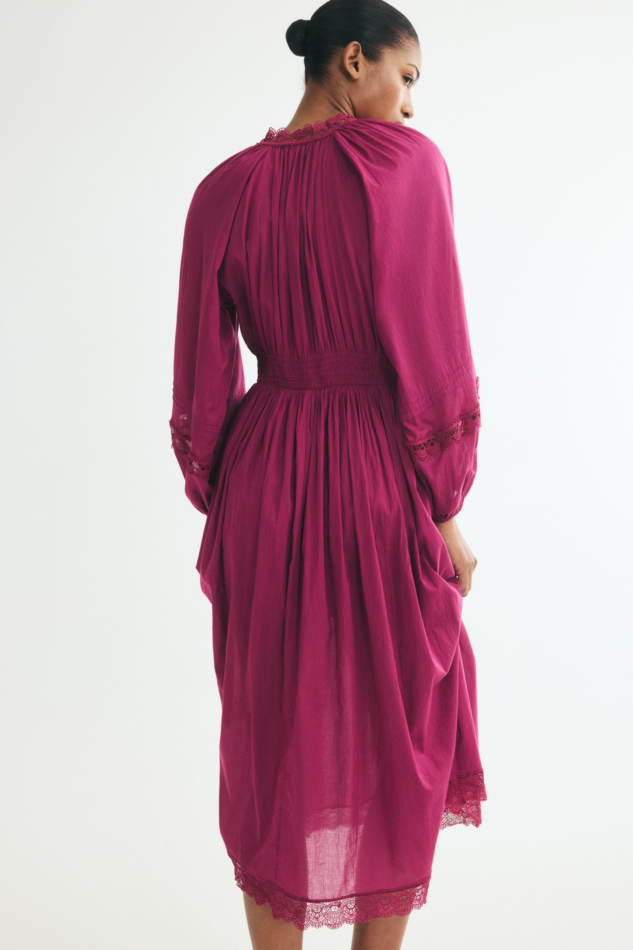 View larger image: A calf-length, magenta cotton dress is shown from the back on a model, showcasing a smocked waist and gathered detailing along the high neckline and skirt. The dress features long balloon sleeves with scalloped lace insets and a flared skirt finished with a scalloped lace hem.