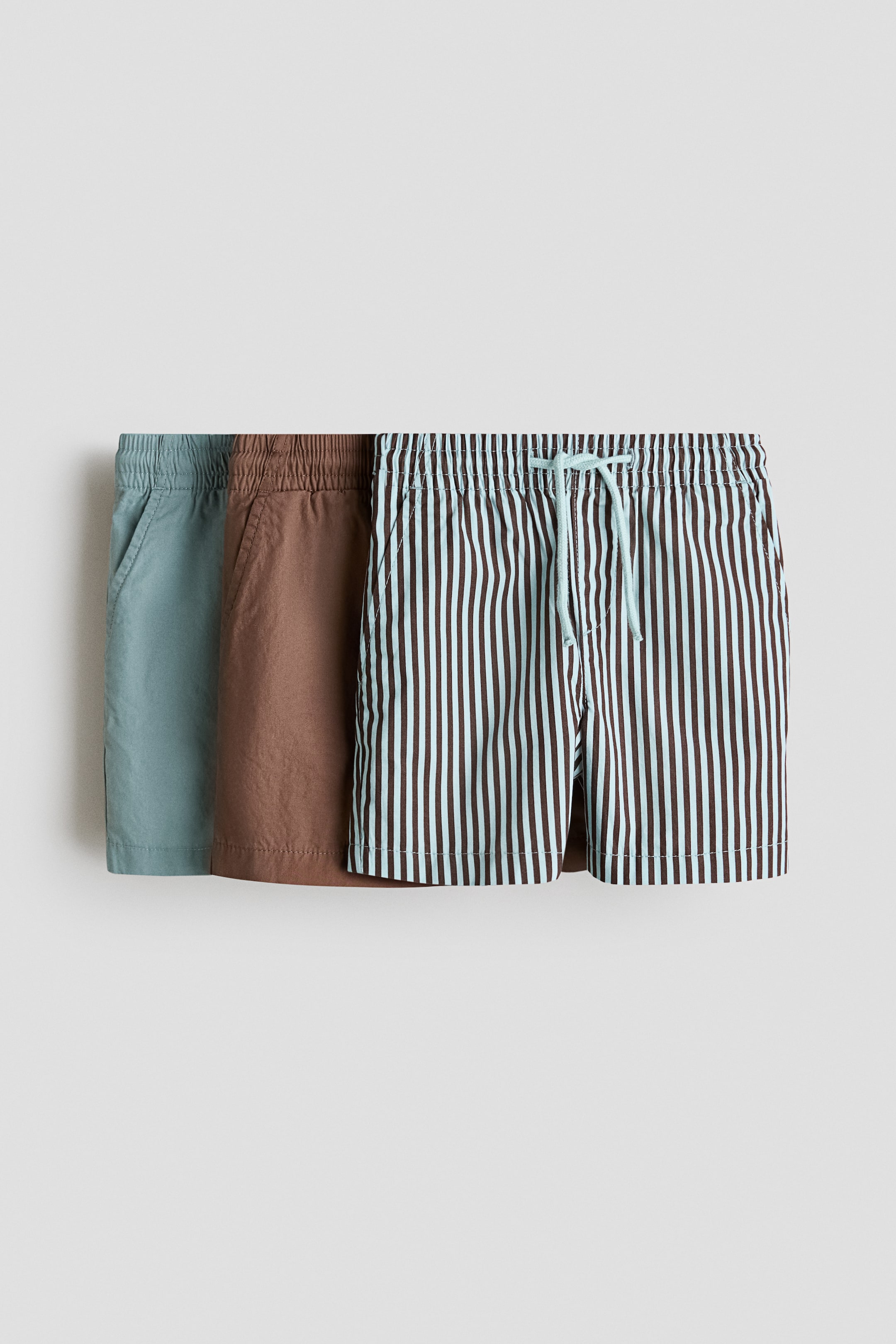 View larger image: Three pairs of crisp cotton poplin shorts are displayed front-facing. Each pair features an elasticated waist and diagonal side pockets. The leftmost pair is solid teal, the middle pair is solid brown, and the rightmost pair is light blue with dark brown vertical stripes, showcasing a light blue drawstring at its waist.