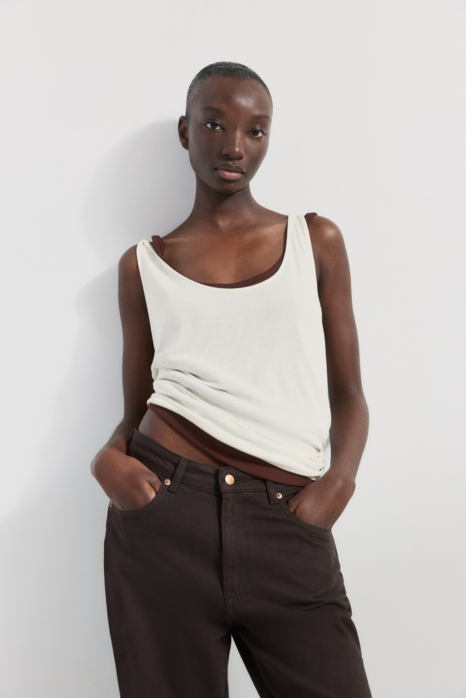 The model is wearing a loose-fit vest top crafted from narrow-ribbed viscose jersey. This cream-coloured top boasts a scoop neckline and deep armholes, layered over a brown top that peeks through at the neckline and hem. The vest top is also partially tucked into dark trousers. - 1