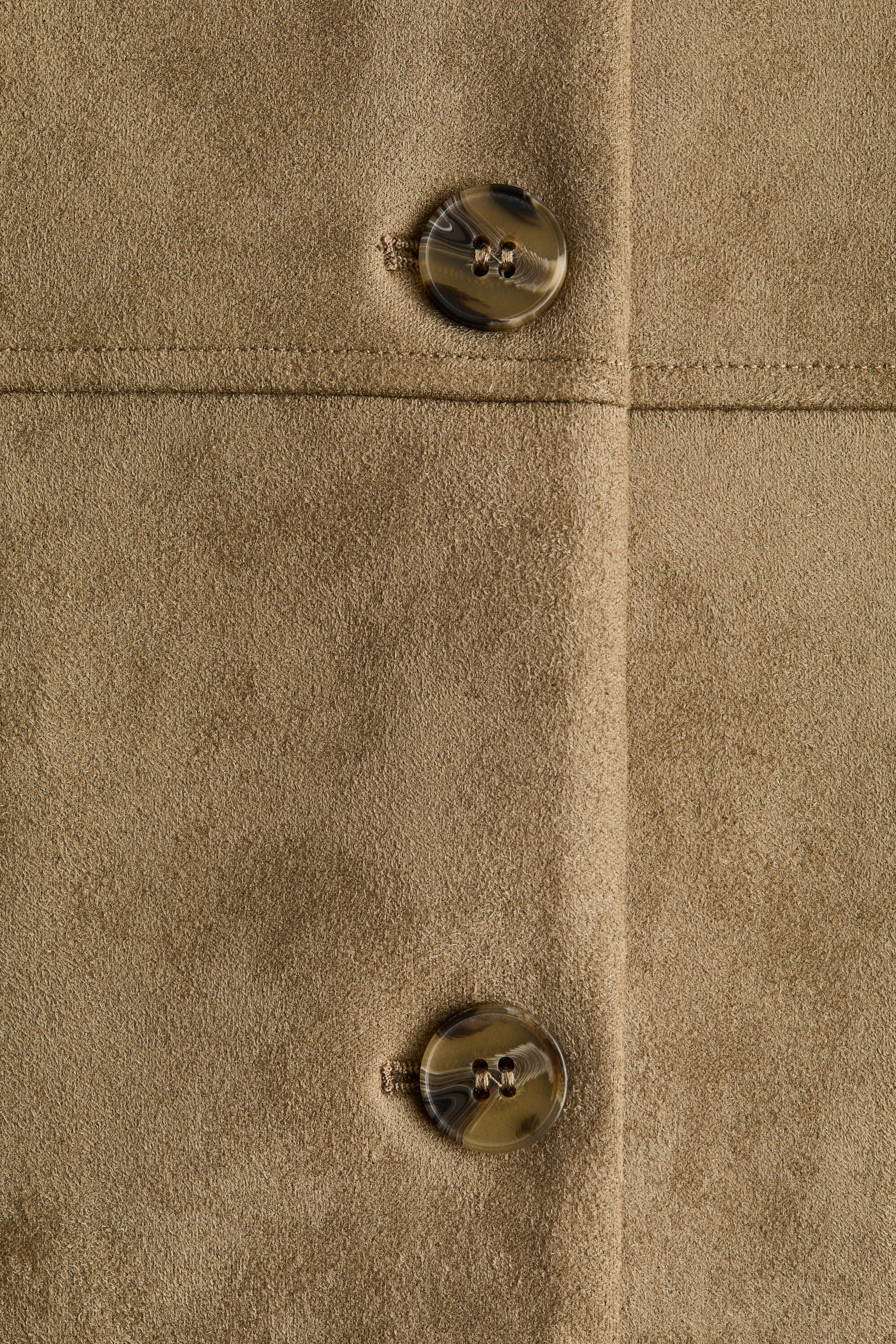 Ampliar la imagen: Una tela de gamuza color beige presenta dos botones redondos de cuatro agujeros con un patrón jaspeado de carey. Una costura horizontal cosida es visible encima del botón superior, y una costura vertical corre a la derecha de los botones.