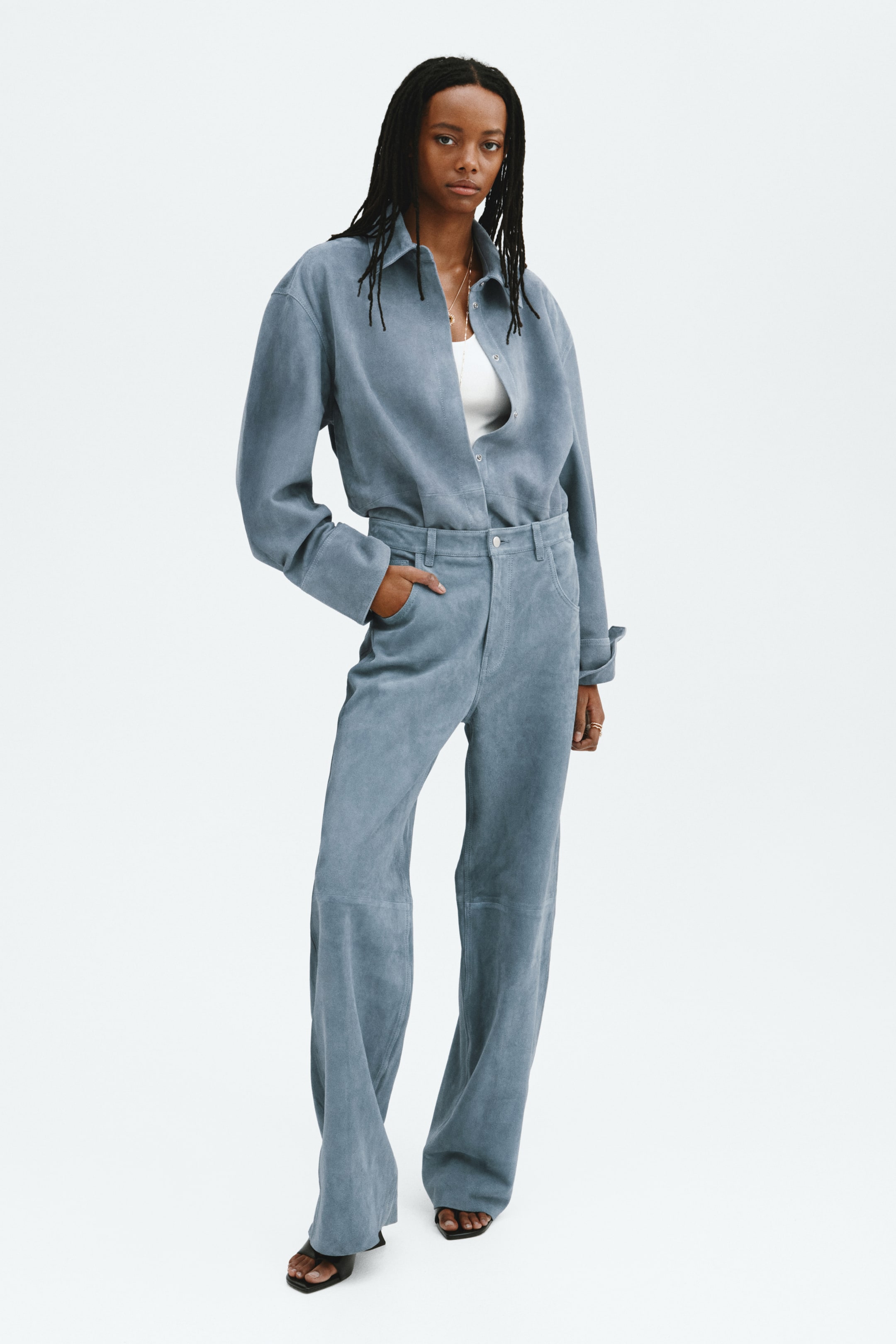 Vezi imaginea mai mare: O cămașă de culoare albastru deschis, asemănătoare cu pielea întoarsă, și pantaloni asortați sunt prezentate dintr-o vedere frontală. Cămașa cu guler, având mâneci lungi cu manșete largi, este stilizată descheiată peste un top alb, în timp ce pantalonii cu talie înaltă sunt proiectați cu o croială largă și detalii vizibile ale cusăturilor.