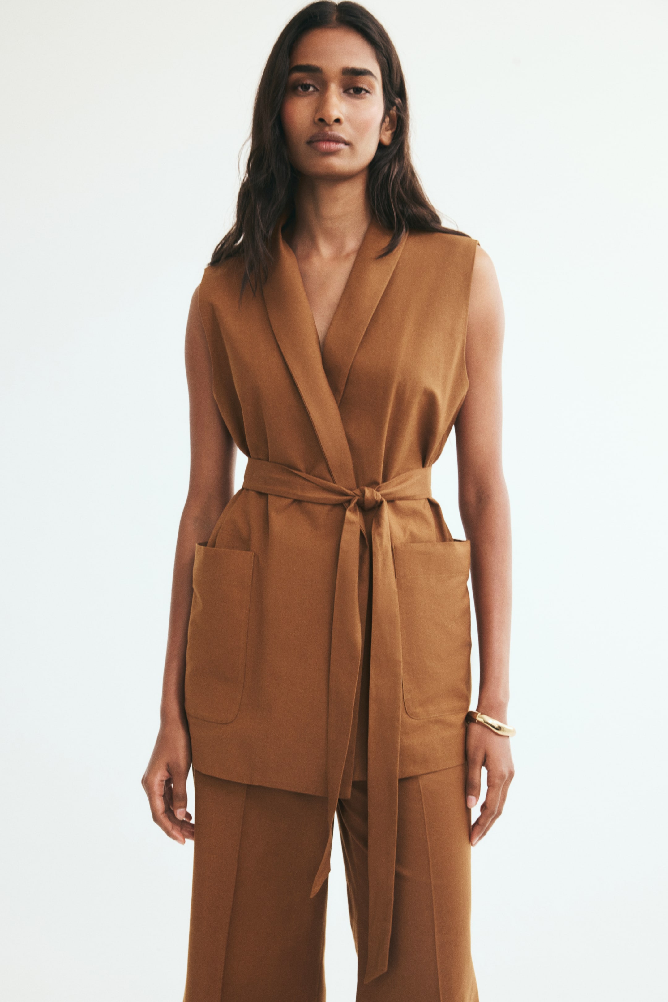 View larger image: A long, brown vest featuring a shawl collar, a wrap front, and open front pockets is worn by the model. The vest is styled with a matching tie belt knotted at the waist and paired with matching trousers.