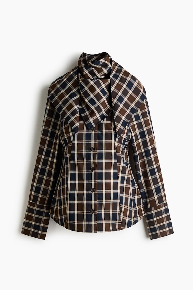 A long-sleeve plaid shirt is facing front, showcasing a brown, navy blue, and white tartan pattern with a button-front closure. It is featuring an exaggerated wide collar layered with a matching draped neck scarf and long sleeves with wide, folded cuffs. - 1