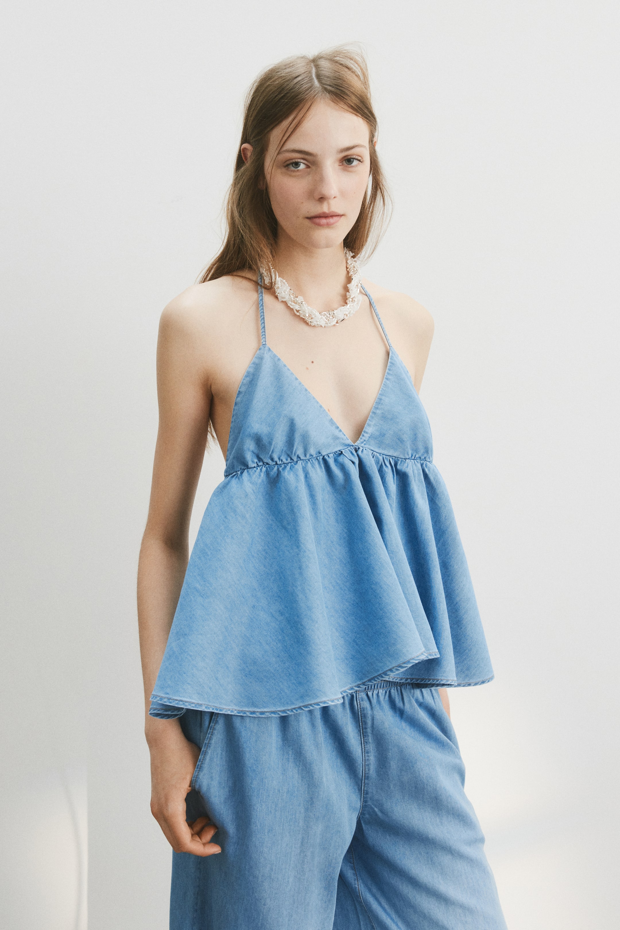 View larger image: A light blue denim halter top features thin straps, a V-neckline, and a gathered peplum hem, flowing over matching relaxed-fit denim pants with an elasticated waistband. The full outfit is displayed from a slight front-right angle.
