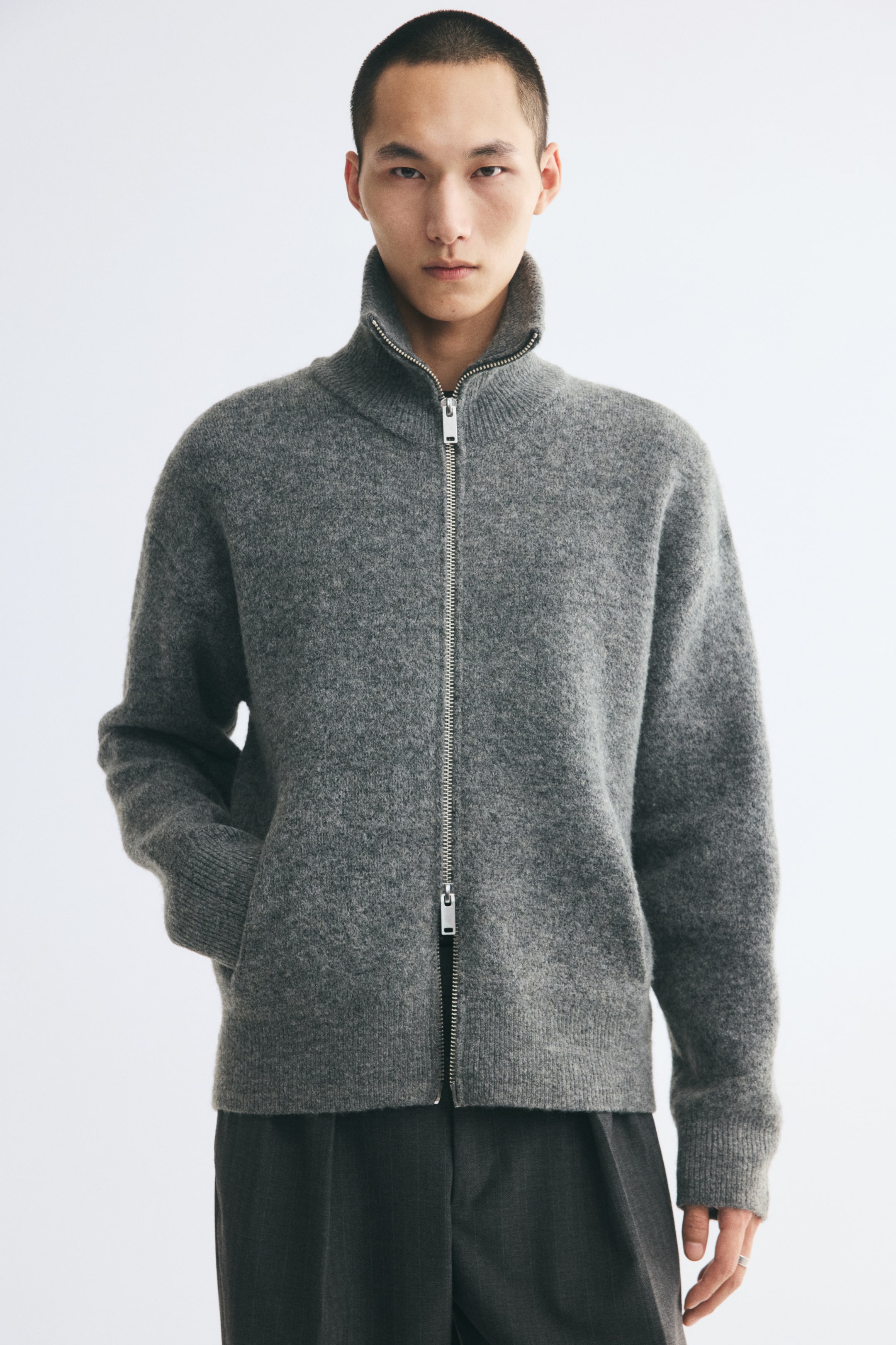 Grösseres Bild ansehen: Eine graue Strickjacke mit gebürsteter Oberfläche wird vom Model getragen, das mit einer Hand in einer Fronttasche steht. Die Strickjacke verfügt über einen gerippten Stehkragen, einen silberfarbenen Zwei-Wege-Reissverschluss vorne, überschnittene Schultern sowie gerippte Abschlüsse an Ärmeln und Saum.