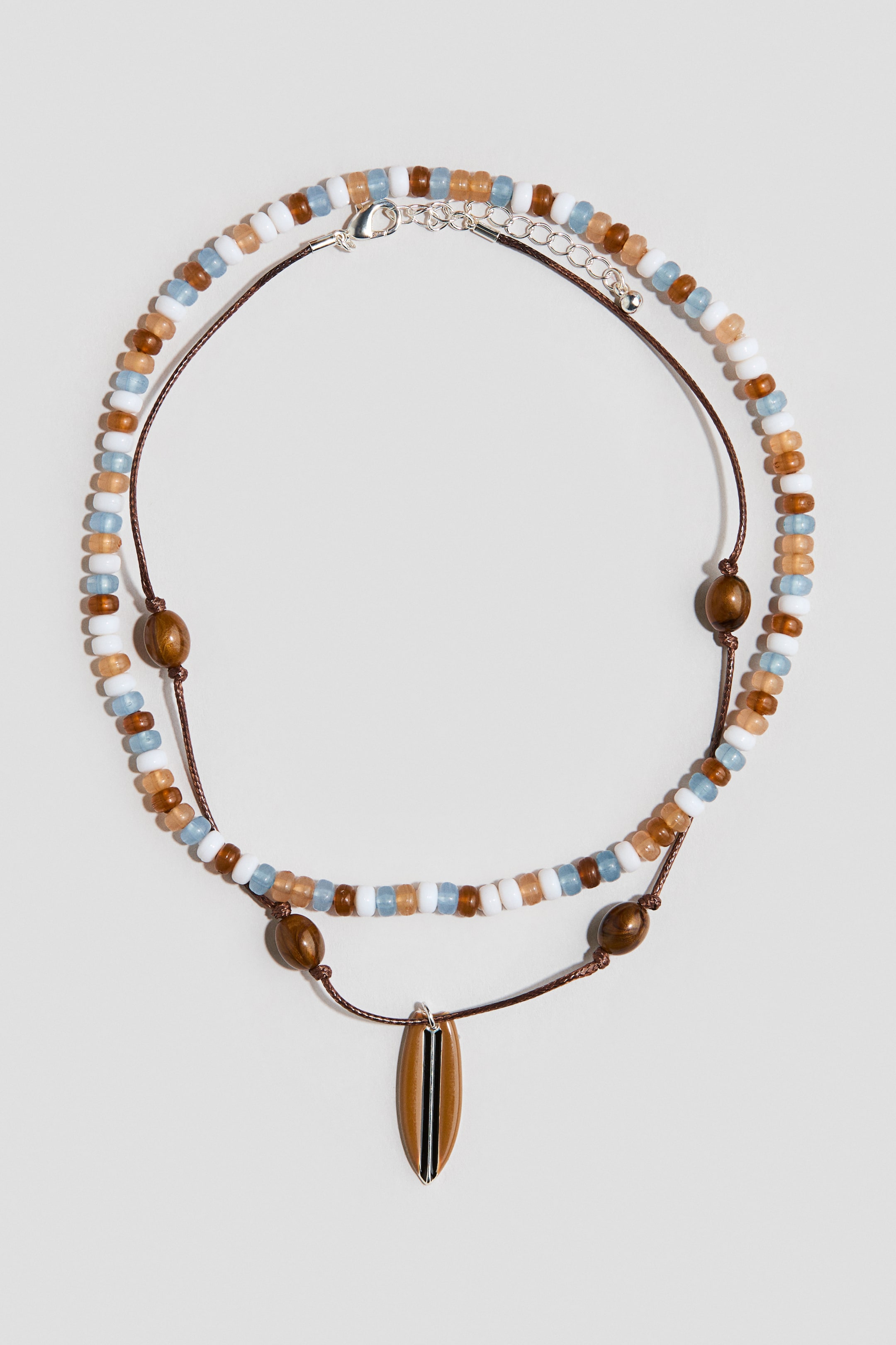 Agrandir l'image: Un collier multi-couches présente un brin de perles en blanc, bleu clair et ambre, ainsi qu'un brin de corde marron avec des perles en bois ovales. Un pendentif en forme de planche de surf marron avec des rayures noires et un sommet argenté est suspendu à la corde, tandis qu'un fermoir mousqueton argenté et une chaîne d'extension sécurisent le brin de perles.