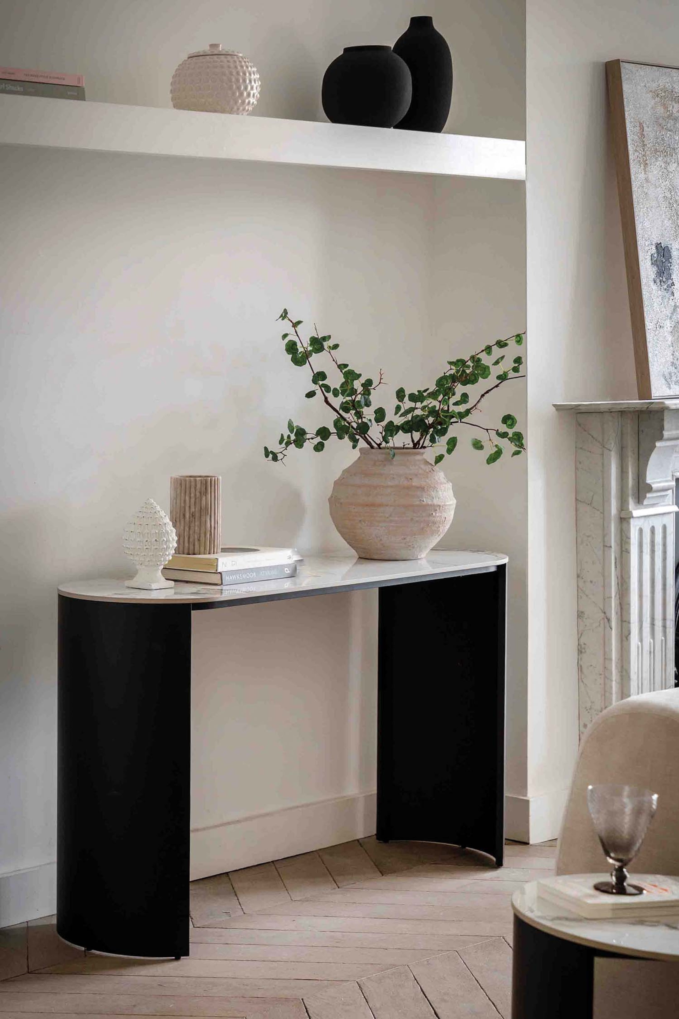 View larger image: A console table featuring a white marble top with subtle grey veining and curved black panel legs. Its design forms a corner, with the top curving to the left and extending straight to the right. Decorative items, including a large textured vase with green branches, are arranged on its surface.