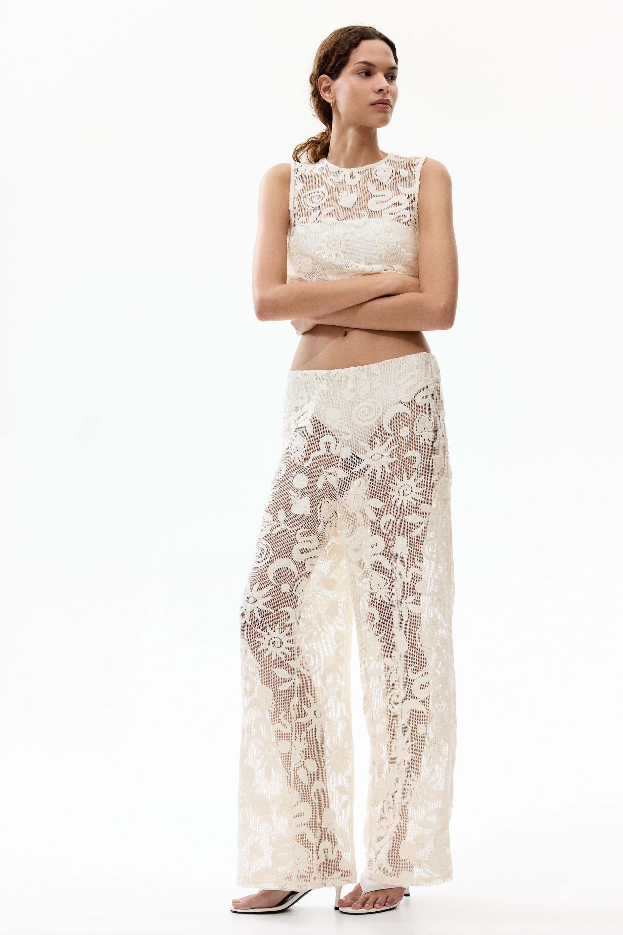 Ver imagem maior: Um conjunto de duas peças em renda de cor creme está a ser apresentado, de frente. O top sem mangas e cortado apresenta um decote redondo e um padrão intrincado de malha aberta, revelando um sutiã leve. As calças compridas e de perna larga a condizer também apresentam o padrão de renda de malha aberta, revelando cuecas leves.