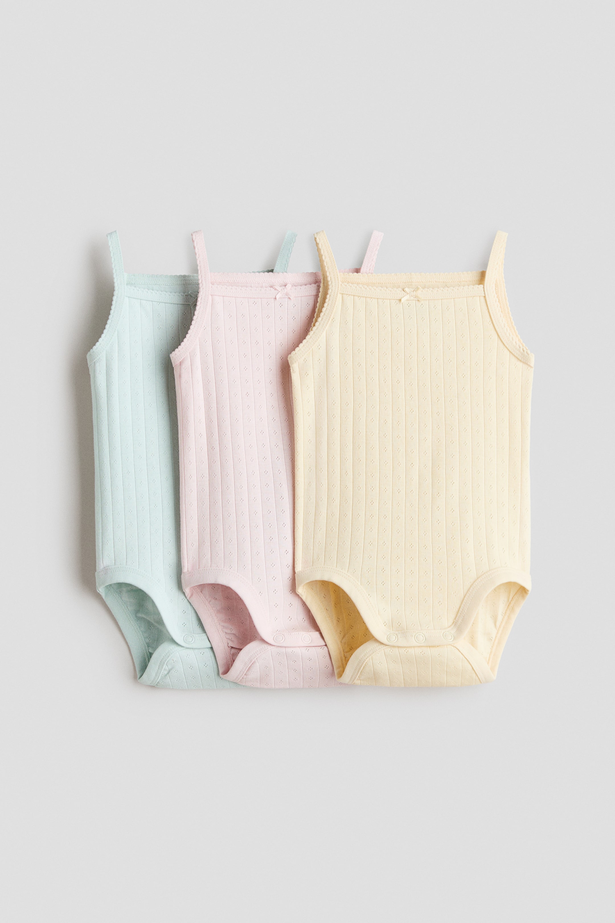 Voir une image plus grande: Trois bodys sans manches sont présentés de face, en bleu clair, rose et jaune. Chaque body est confectionné en jersey de coton côtelé et perforé, avec de fines bretelles ornées de petits nœuds et des boutons-pression à l'entrejambe.
