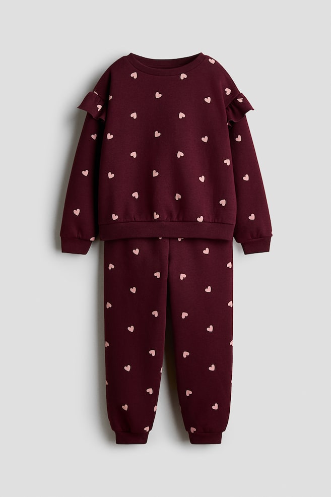 A front view of a two-piece sweater and track pants set in burgundy fleece featuring an all-over pink heart pattern. The long-sleeved sweater is showing a ribbed round neckline, dropped shoulders with flounce trims, and ribbed cuffs and hem, paired with matching track pants featuring an elasticated waistband and ribbed ankle cuffs. - 1