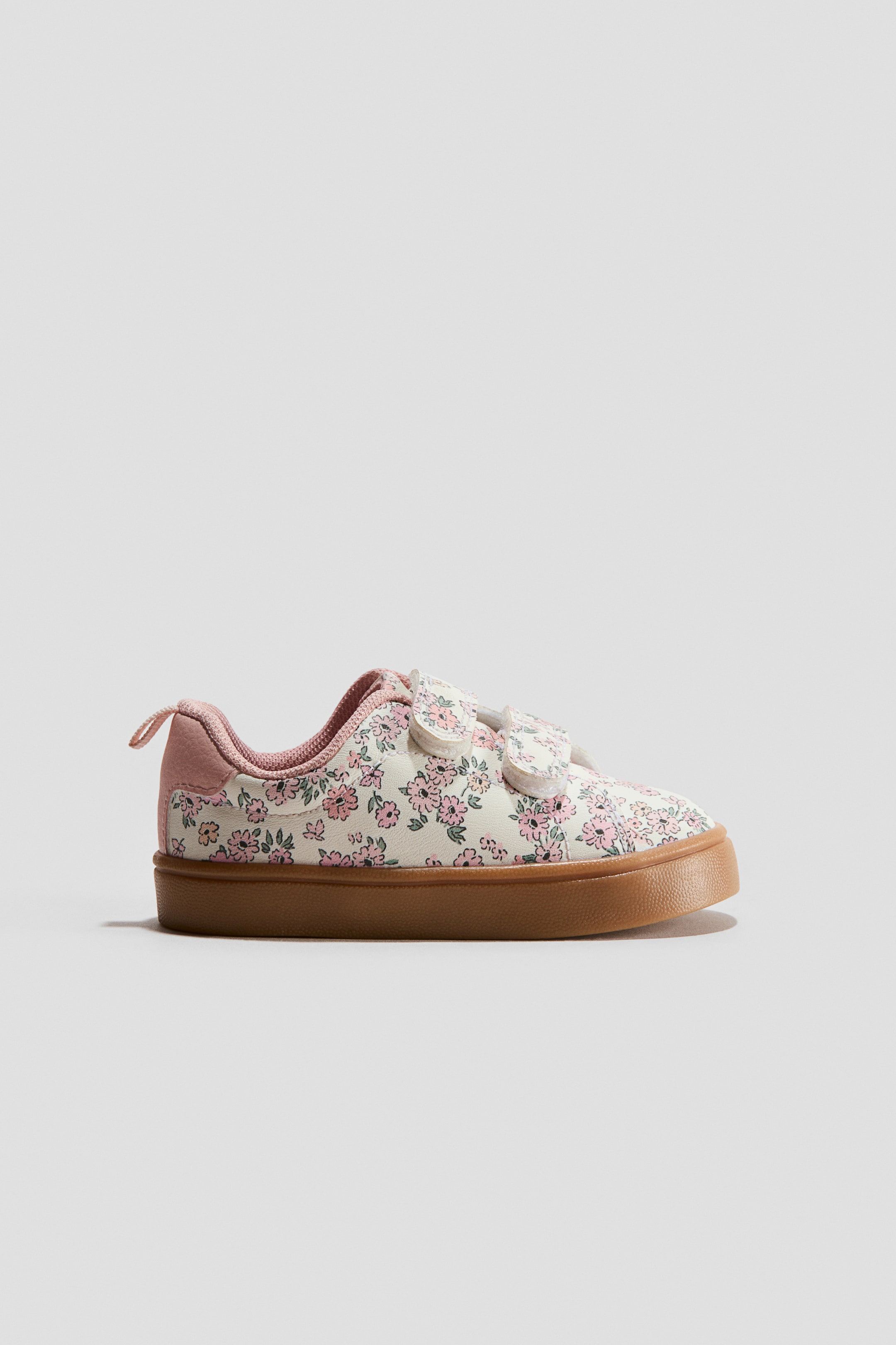 View larger image: A sneaker is facing right, with a cream upper featuring a pink and green floral pattern. Two white hook-and-loop straps secure the instep, while a solid pink heel counter, collar lining, and pink pull tab detail the heel, and a light brown rubber sole completes the shoe.
