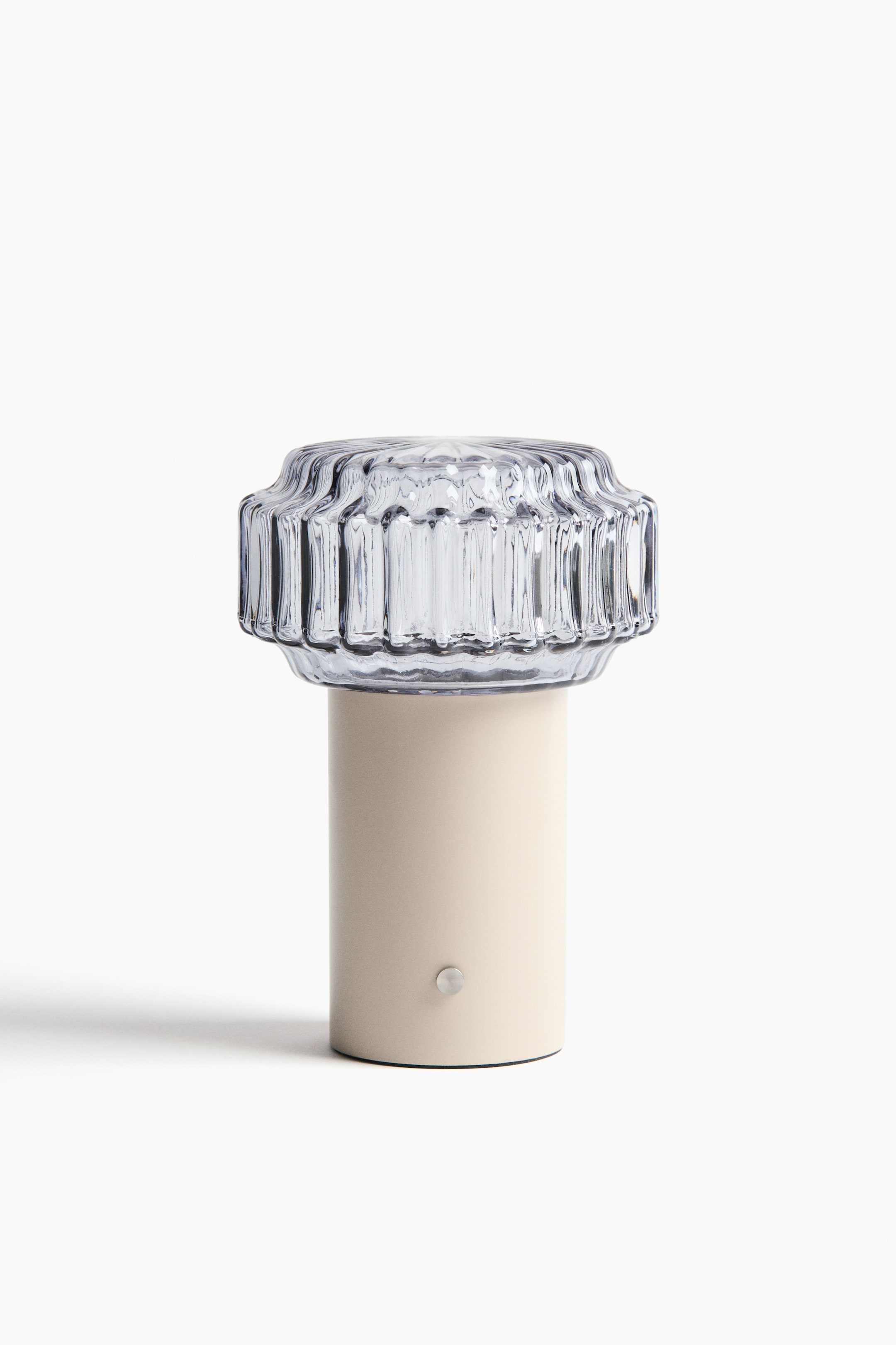 View larger image: A table lamp is featuring a cream-colored, cylindrical base with a small, round, silver button visible on its front, and a clear, fluted glass shade is resting on top.