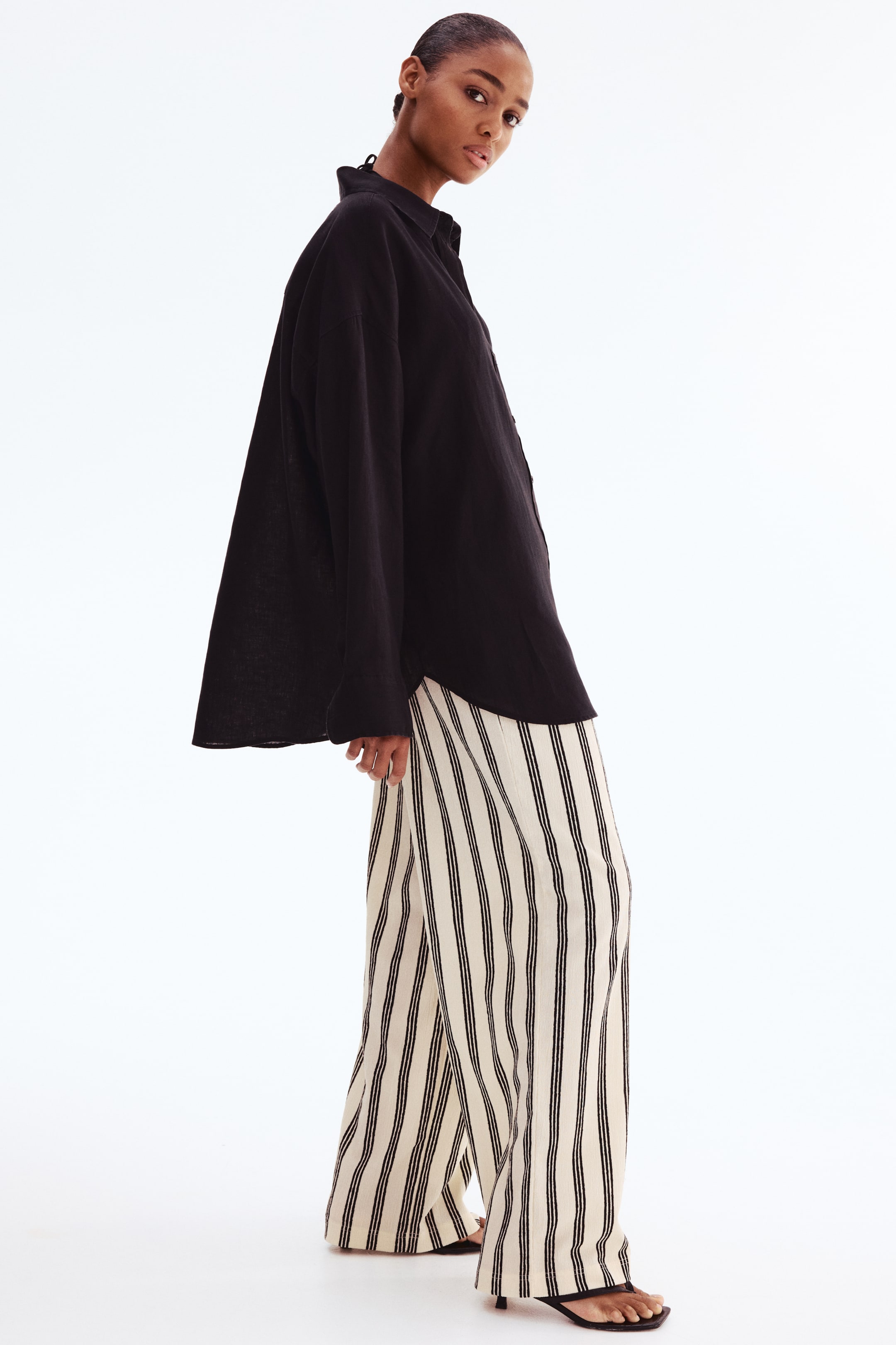 View larger image: From a side view, a black oversized button-down shirt features a pointed collar and long, loose sleeves. It is paired with cream-colored wide-leg pants that feature vertical black stripes and drape loosely to the ankles.