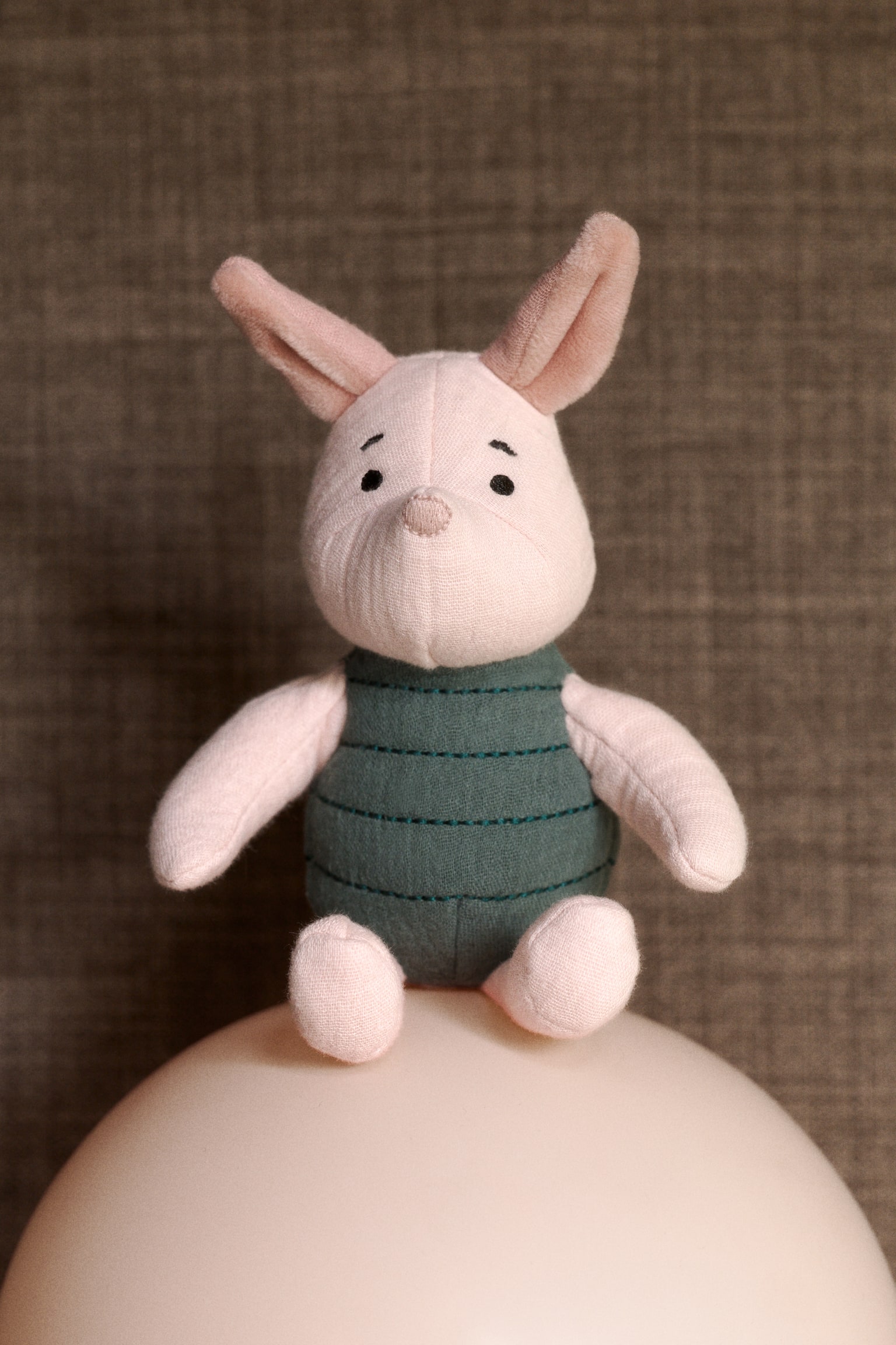 A soft cotton muslin rattle, shaped as a character, is facing front. It is featuring a light pink head with embroidered black eyes and a pink nose, and long pink ears. Its body is dark green with horizontal stitched lines. - 1
