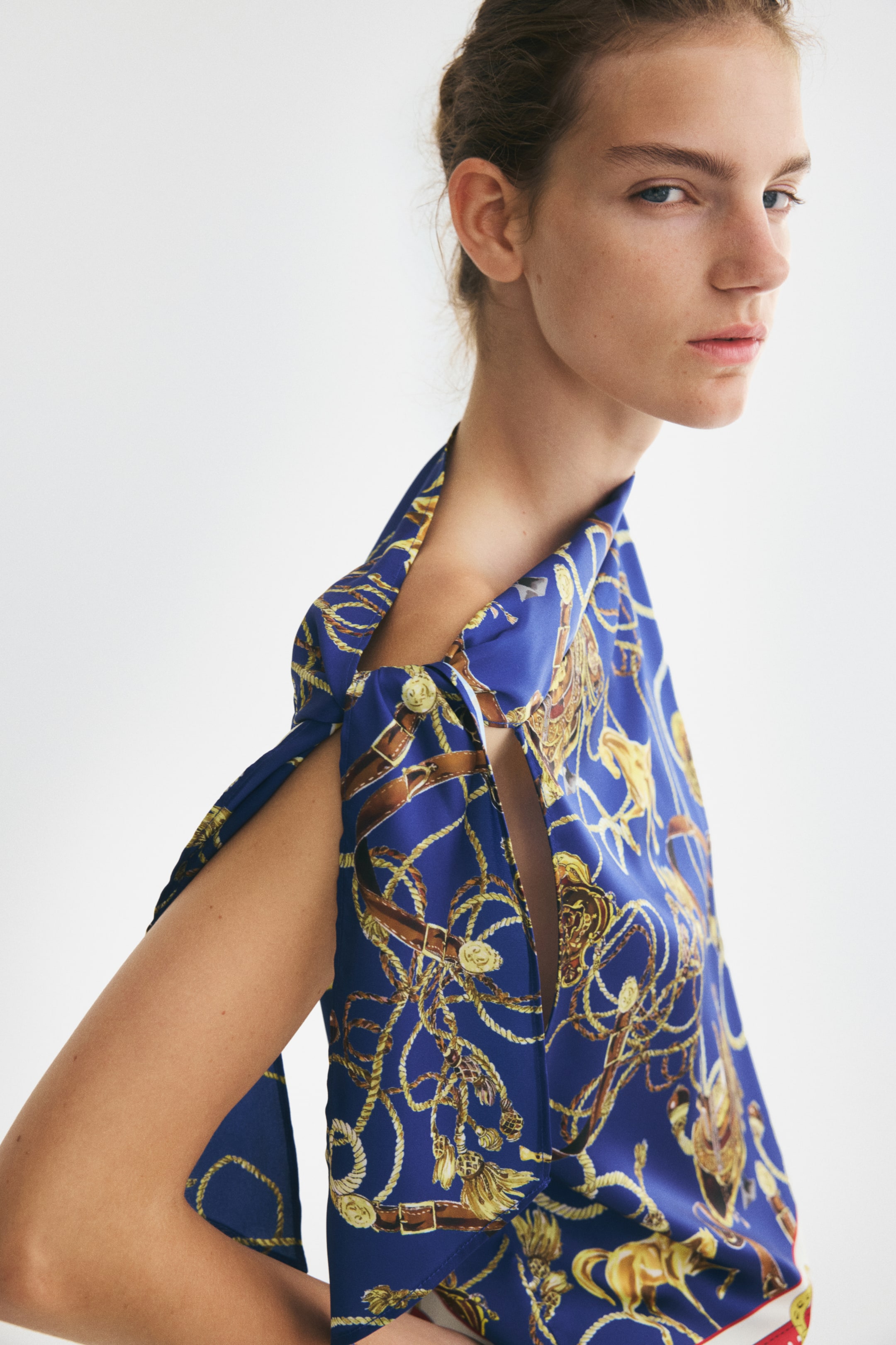 Agrandir l'image: Un haut en satin sergé bleu, orné d’un motif équestre composé de cordes dorées et de ceintures marron, est porté par le mannequin. Le vêtement présente un décolleté asymétrique avec un détail drapé rabattu et une ouverture découpée à l’épaule.