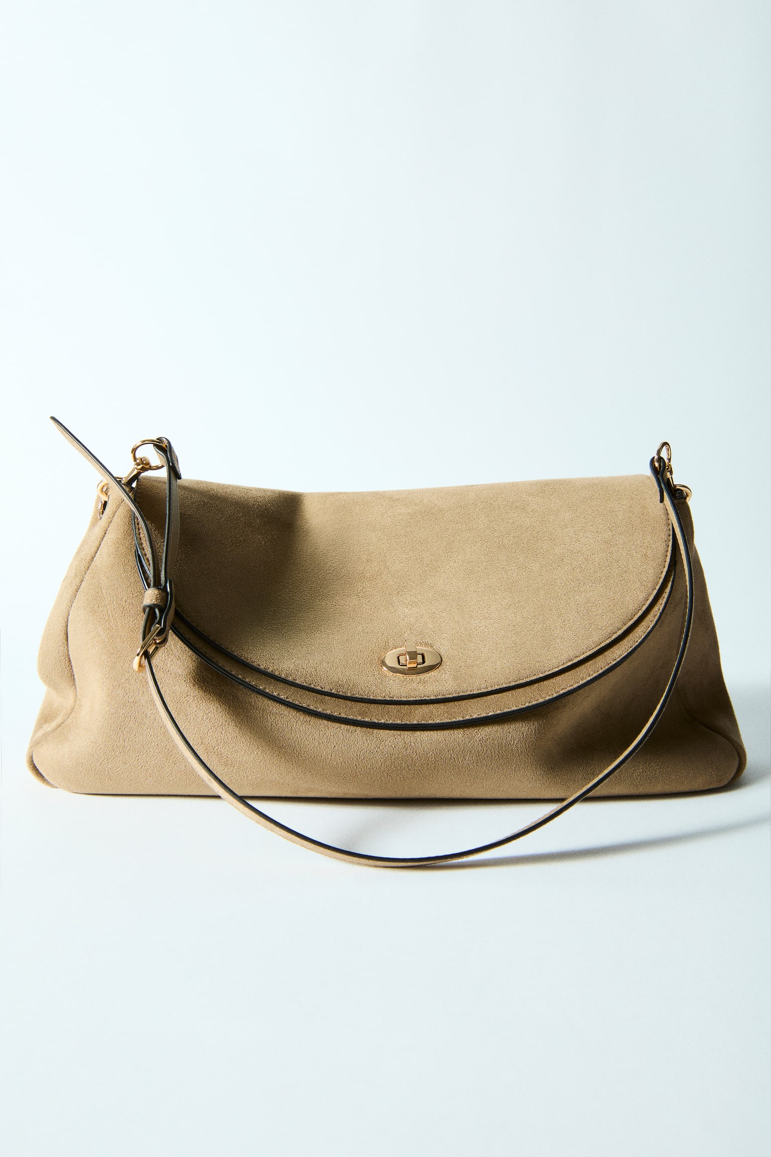 A taupe suede shoulder bag is positioned facing forward, showcasing a curved flap closure with dark piping, fastened by a gold-toned turn-lock. An adjustable shoulder strap extends upwards from its sides. - 1