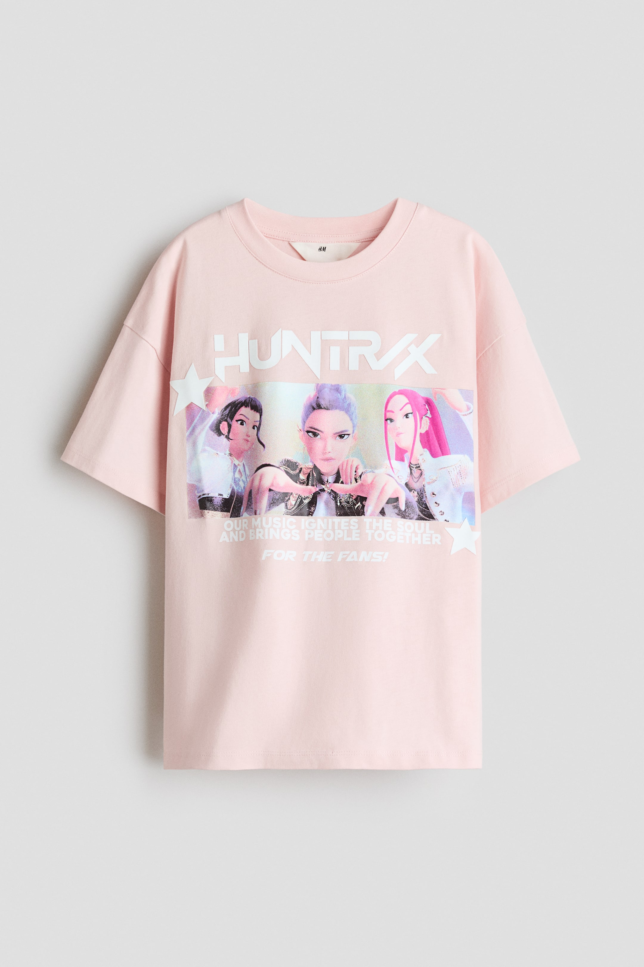 Visualizza immagine più grande: Una t-shirt oversize in morbido jersey di cotone rosa chiaro presenta il suo fronte, caratterizzato da un girocollo con bordo a coste e spalle cadenti. Sul davanti è presente una grande stampa grafica con "HUNTRX" in lettere bianche e grassetto sopra tre personaggi animati femminili, accompagnati da testo bianco e stelle.