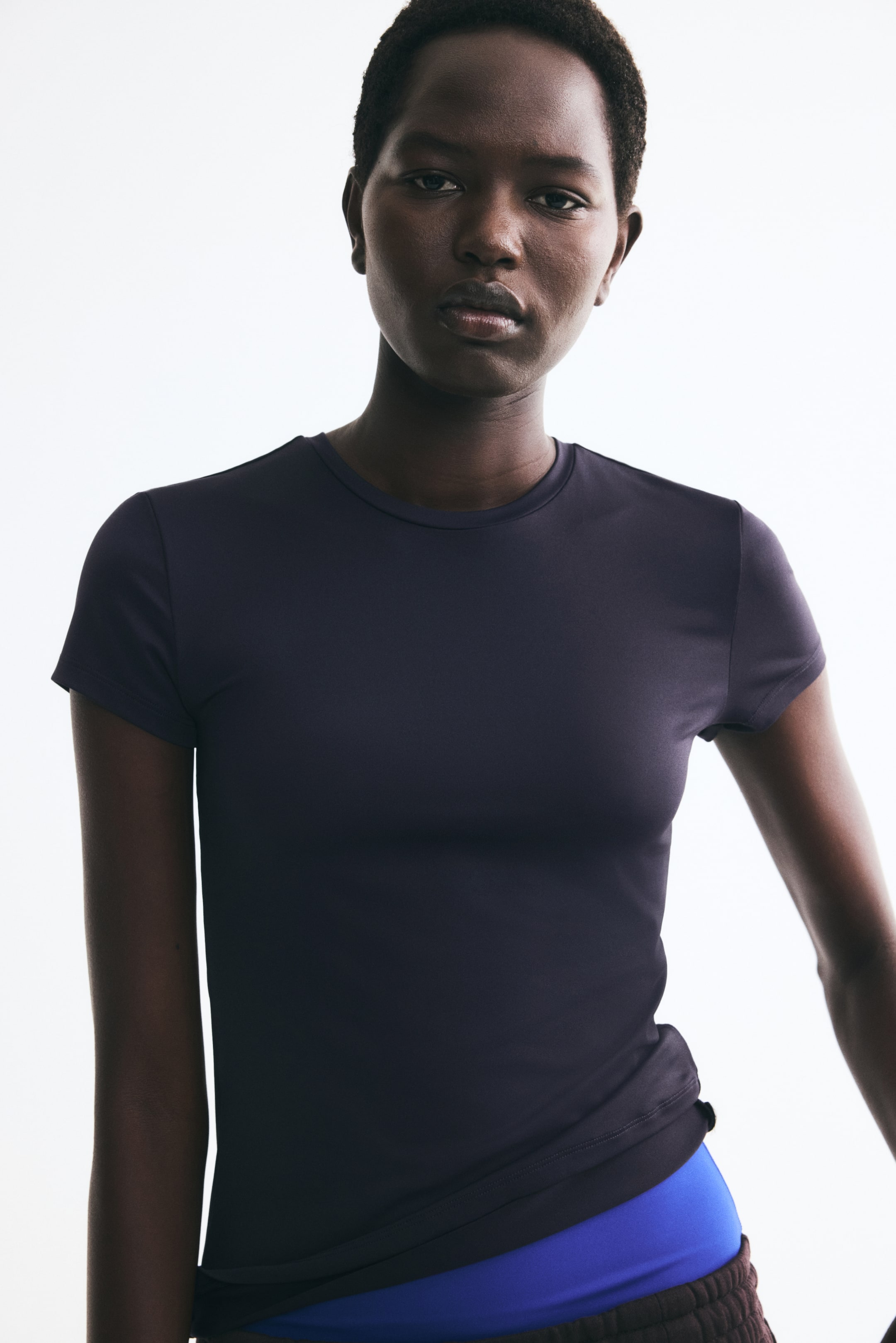 View larger image: A fitted microfibre tee in dark purple is being worn by the model, featuring short sleeves and a narrow trim around the crew neckline. The hem of the tee is layered over a bright blue waistband.