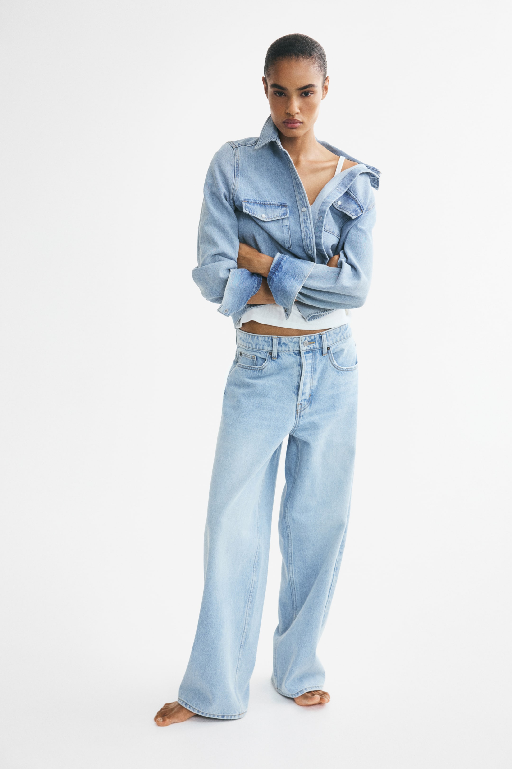 View larger image: The model is wearing a light wash denim shirt jacket featuring chest pockets, styled open over a white crop top with one side elegantly draped off the shoulder. Complementing the look, the model dons light wash wide-leg jeans with a high waist and button closure.