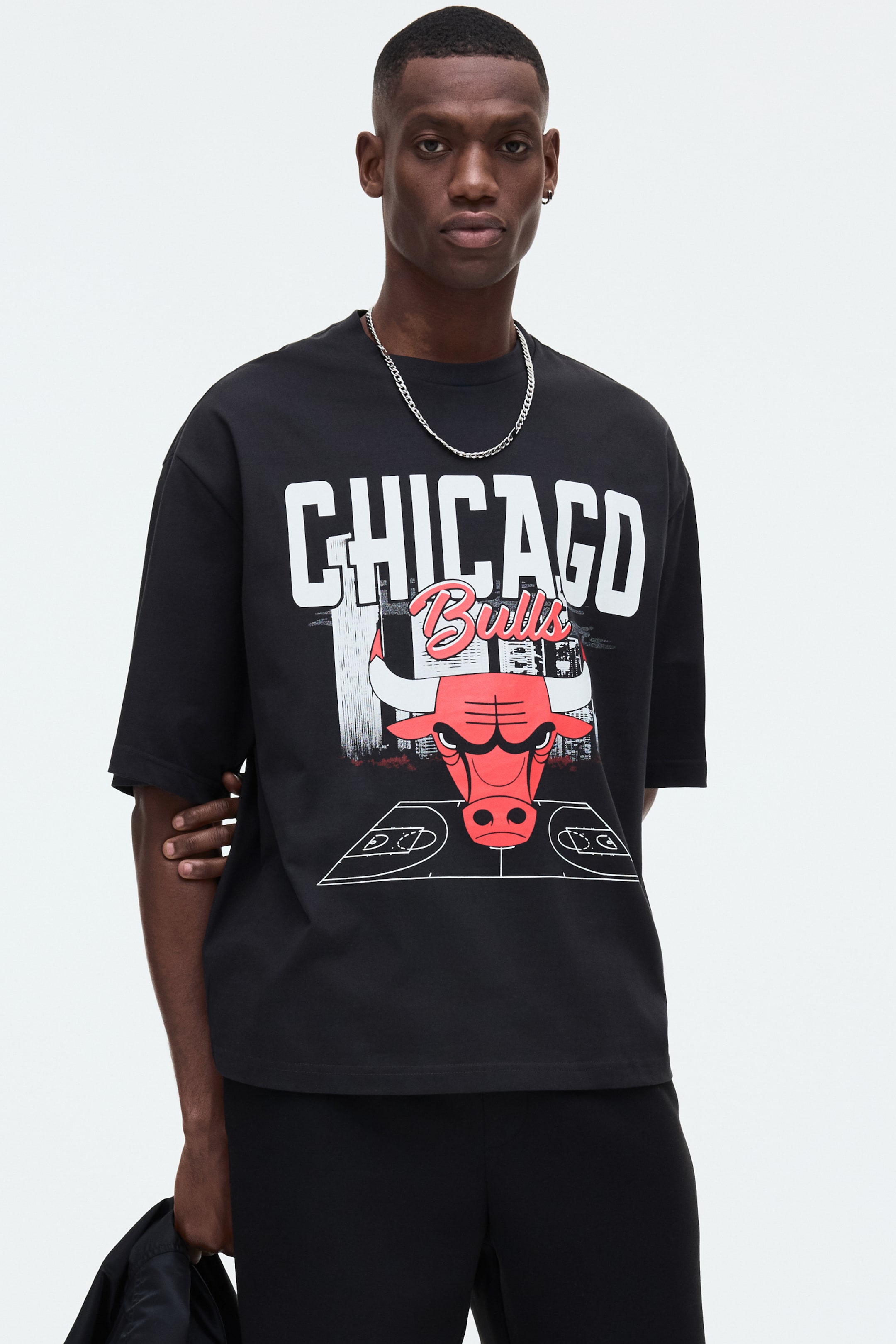 Visa större bild: En svart oversized t-shirt med en stor frontgrafik som visar "CHICAGO Bulls" i vita blockbokstäver och röd skrivstil, med en röd tjurhuvudlogotyp och en basketplan i kontur. Den är designad med en rund halsringning och korta, lösa ärmar.
