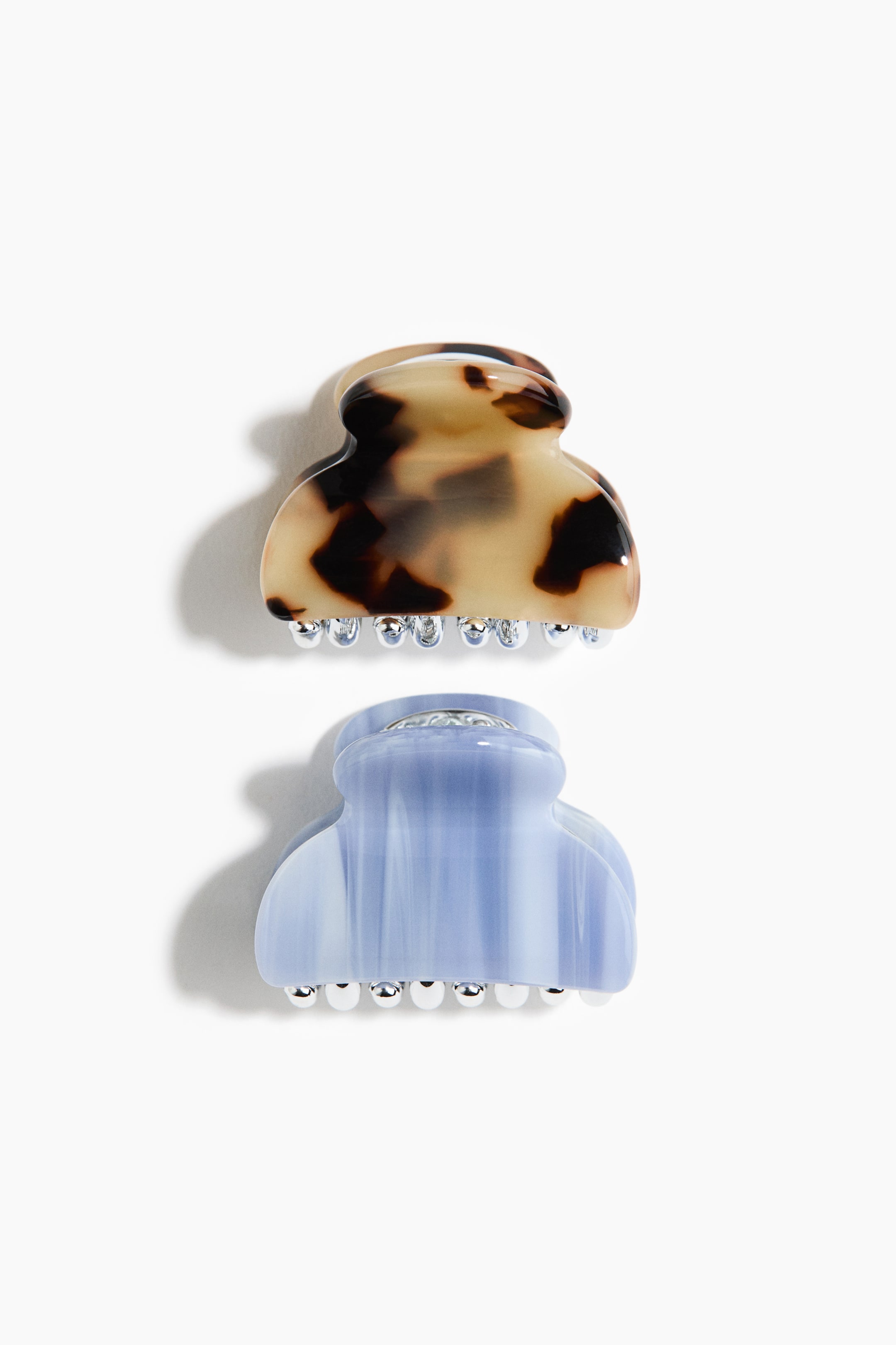 Ver imagen más grande: Se muestran dos pinzas tipo garra para el cabello, vistas desde un ángulo ligeramente elevado. La pinza superior presenta un diseño carey en tonos marrón y crema, mientras que la pinza inferior muestra un azul claro translúcido con vetas blancas. Ambas pinzas exhiben dientes redondeados de color plateado a lo largo de sus bordes inferiores.