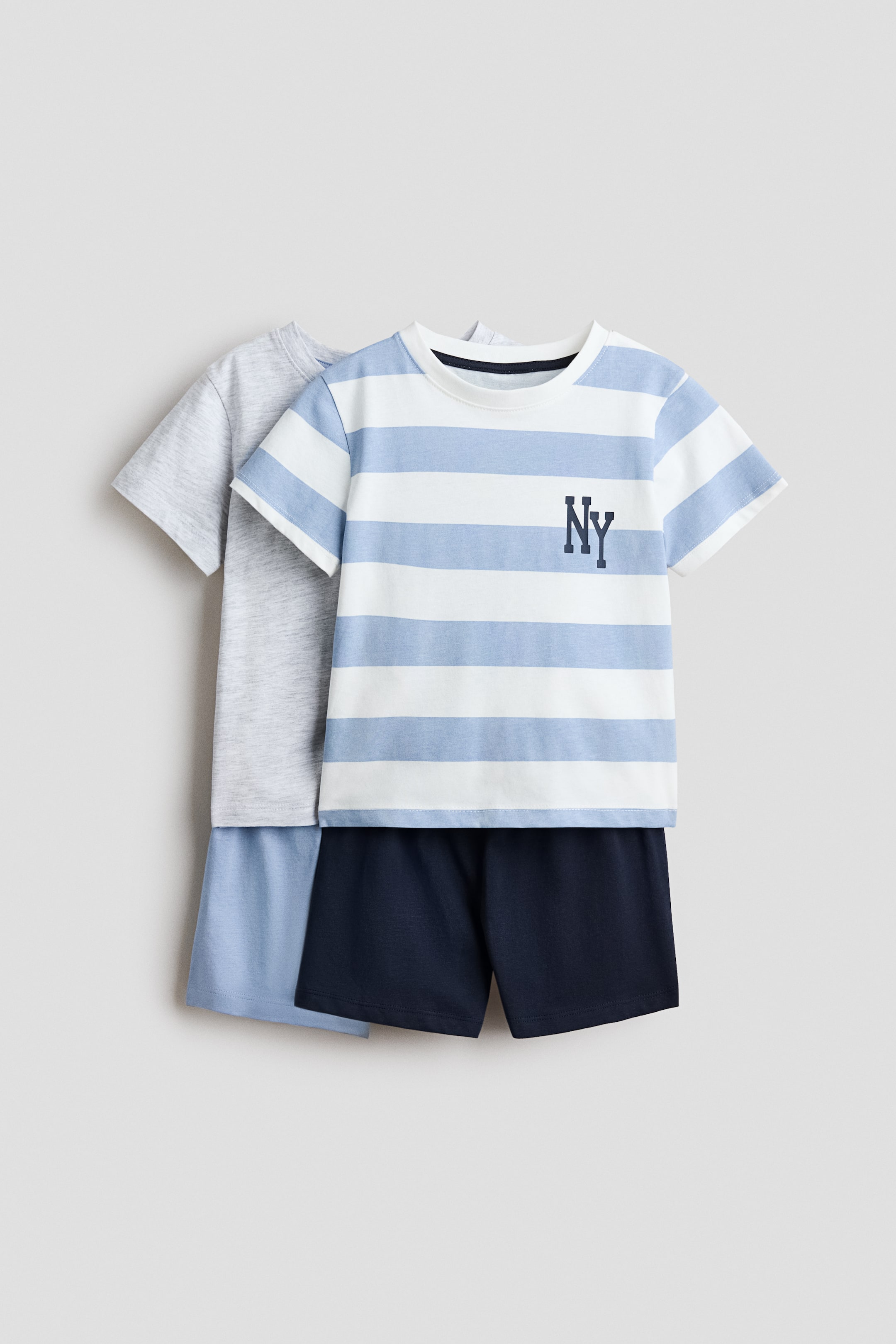 View larger image: A two-pack of pyjama sets is displayed, with one set partially overlapping the other. The front set features a white and light blue striped t-shirt with a dark blue 'NY' logo on the chest, a ribbed neckline, and gently dropped shoulders, paired with dark blue shorts. The second set, visible behind, includes a plain light grey t-shirt and light blue shorts.