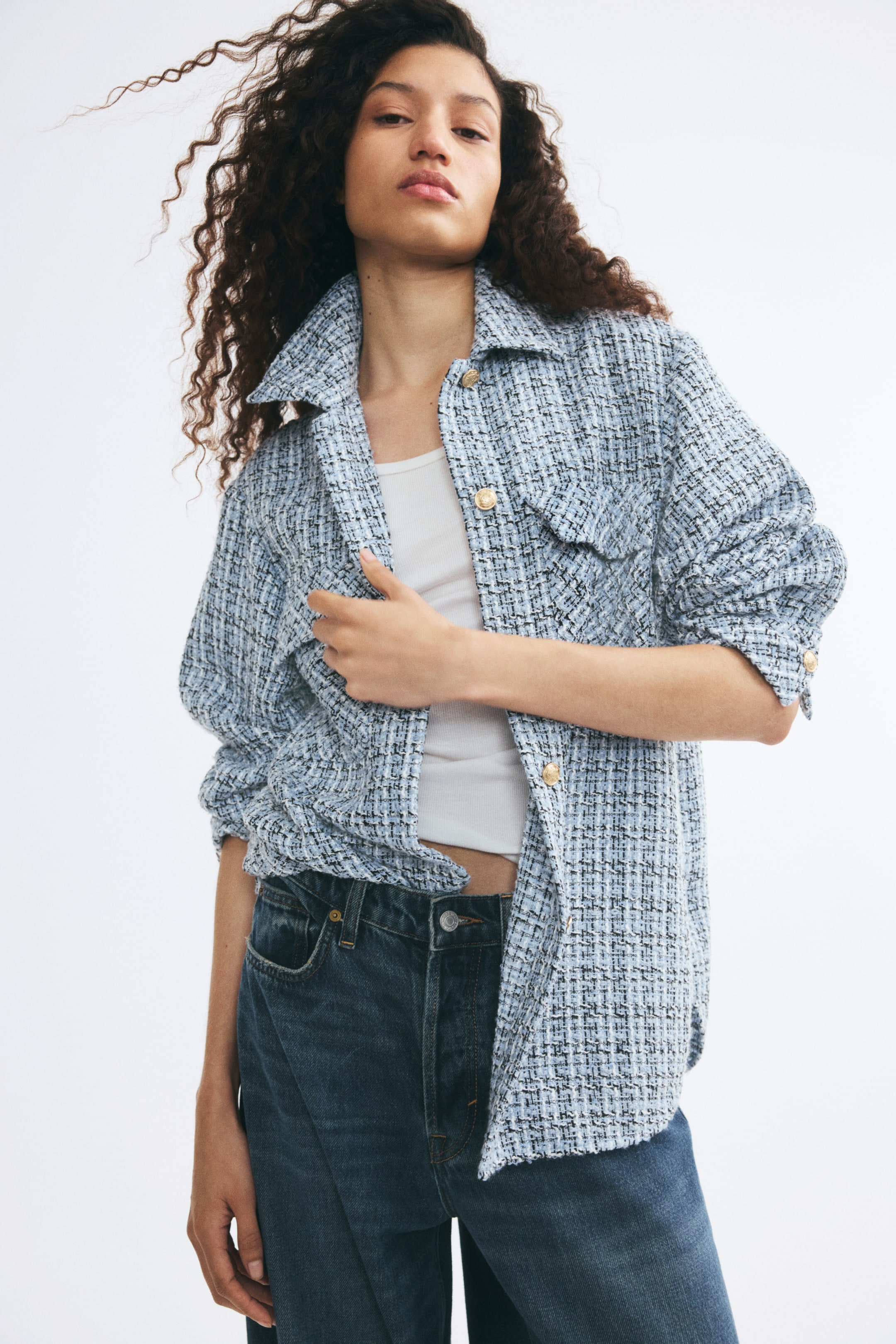 View larger image: The model is wearing a light blue textured tweed shacket open over a white tank top and dark-wash jeans, accented with gold-tone buttons. She is holding one side of the jacket’s front opening, highlighting its relaxed fit and woven fabric.